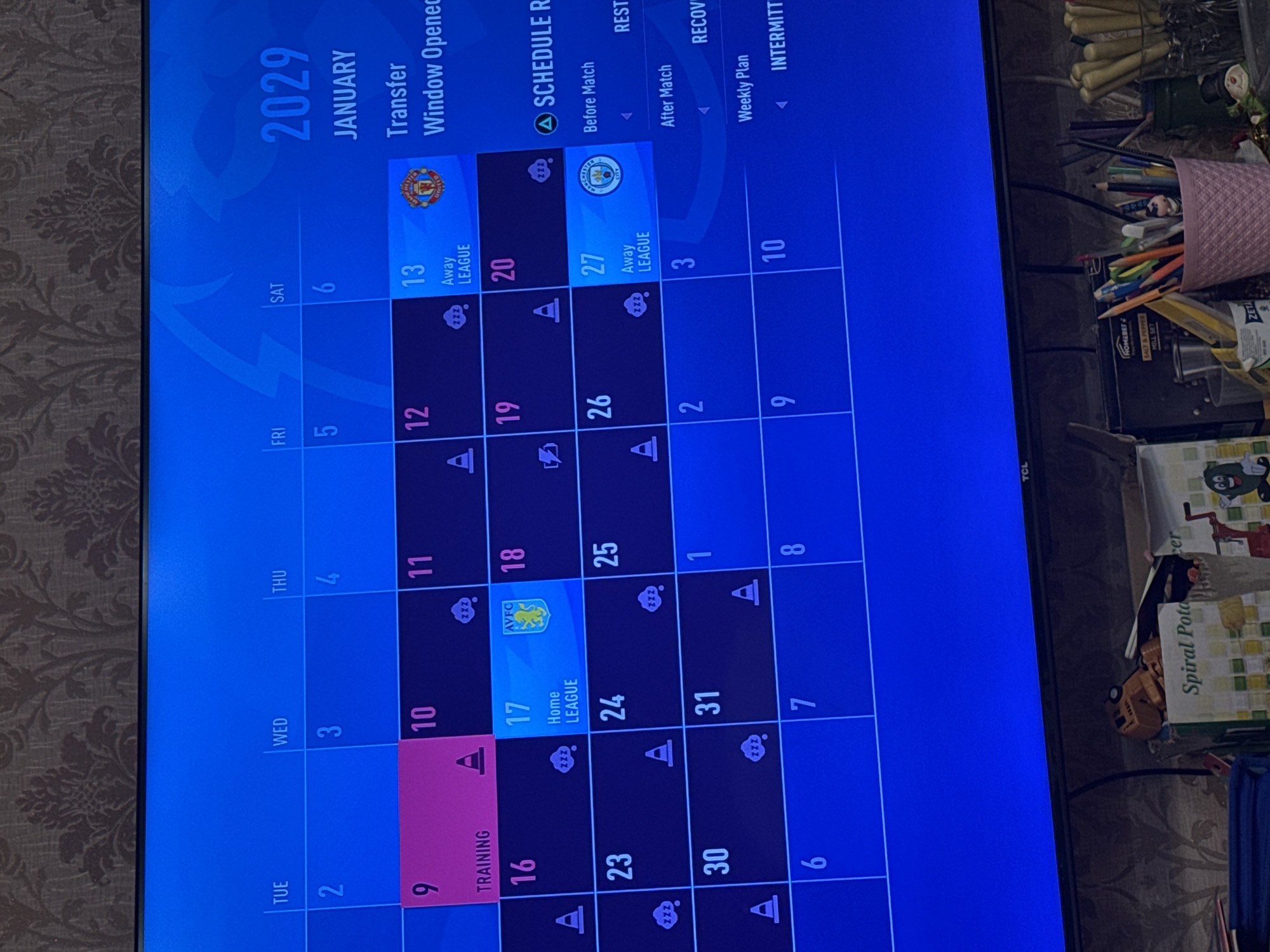 The image shows a television screen displaying a digital calendar for January 2029. The screen indicates that it is a "Transfer Window" and shows a football schedule with training days and away/home league matches against teams such as Manchester United, Aston Villa, and Manchester City. The calendar also displays weekly plan options for players, such as rest and recovery. The surrounding environment visible in the image contains various objects, including a pencil holder with pencils and pens, a salt and pepper mill set, and miscellaneous items.