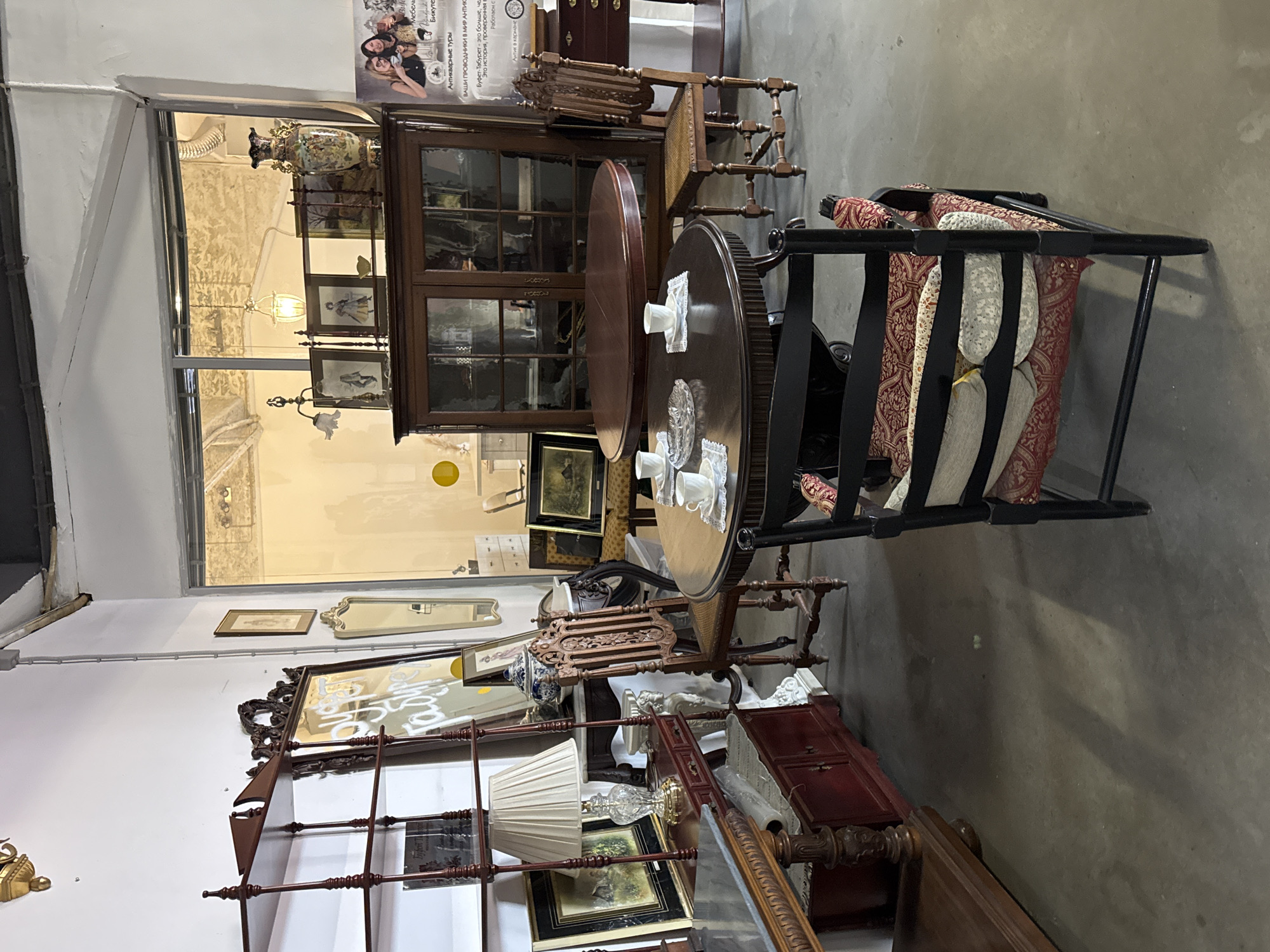 Here is a forensic description of the image:
**Location:** The interior of what appears to be an antique store, presumably in Saint Petersburg, Russia based on the text on the poster.
**Subjects:** The store is filled with antique furniture and decorative objects. There are chairs, tables, cabinets, mirrors, lamps, and framed pictures on display.
**Objects:**
- Antique wooden furniture: Tables, chairs, cabinets, and shelving units.
- Mirrors with ornate frames.
- Table lamps with decorative bases and shades.
- Framed pictures and artwork.
- Decorative items such as vases and porcelain.
- A poster with text in Russian.
**Environment:** The store has a window that offers a glimpse of the outside, but it mainly focuses on the cluttered interior space filled with various antique items. The floor is concrete, and the walls are painted white.
**Details:** The arrangement of objects suggests a slightly disorganized but densely packed showroom style. The lighting appears to be a mix of natural and artificial. The poster on the wall contains text and images in Russian, likely advertising the store or antique tours.