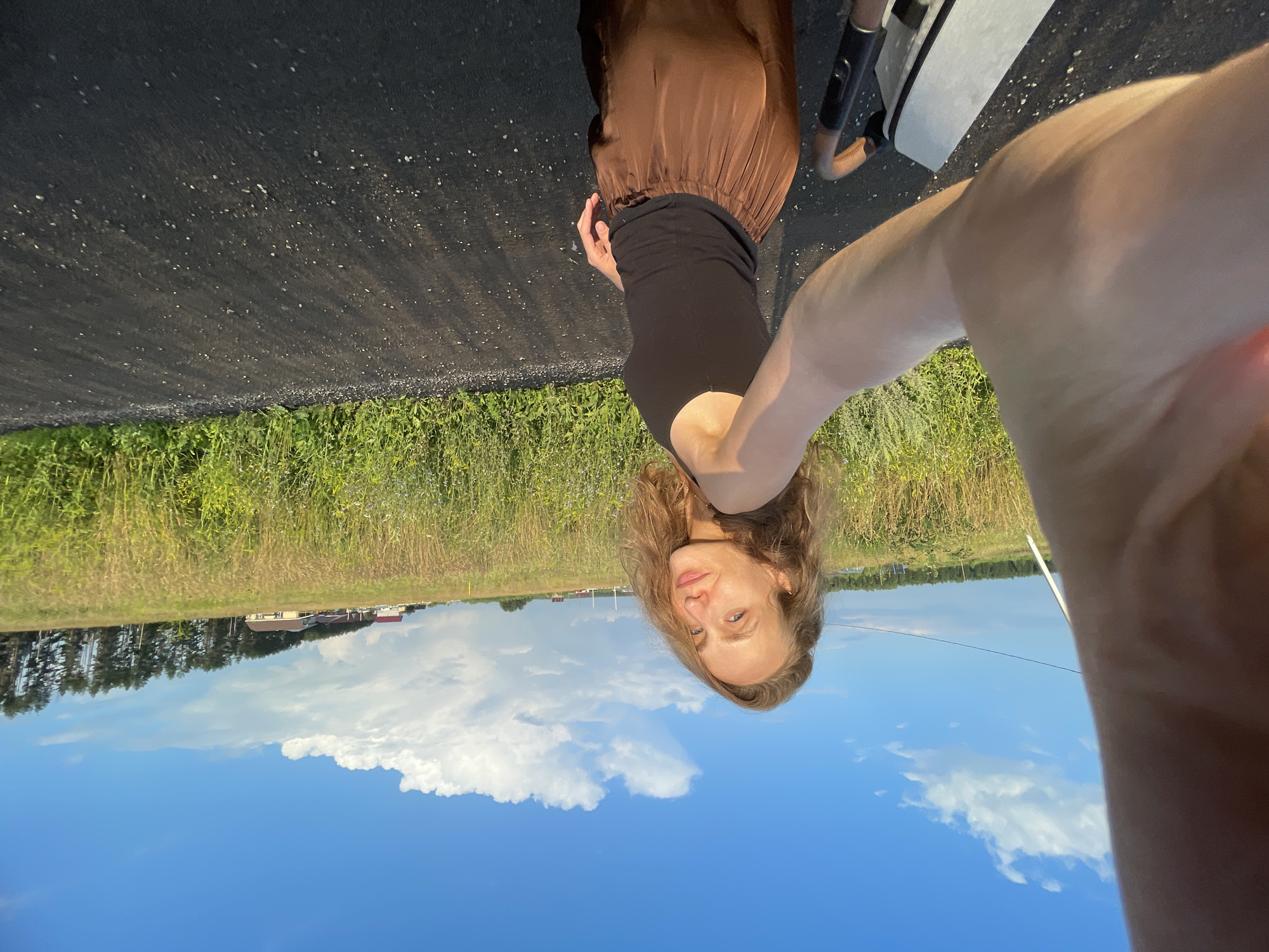 A young woman with long, wavy blonde hair is captured in a selfie-style photograph. She is wearing a black tank top and a flowing, chocolate-brown satin skirt. The woman is positioned outdoors, standing on an asphalt road. Behind her, a field of dry, yellow grass stretches towards a line of trees and a few scattered houses, indicating a rural or suburban setting. The sky above is a vibrant blue, dotted with fluffy white clouds. The lighting suggests it is late afternoon or early evening. The presence of a stroller handle and frame on the left side of the image indicates the woman is likely pushing or accompanying a stroller. The specific location is Podshivalovo, Russia.