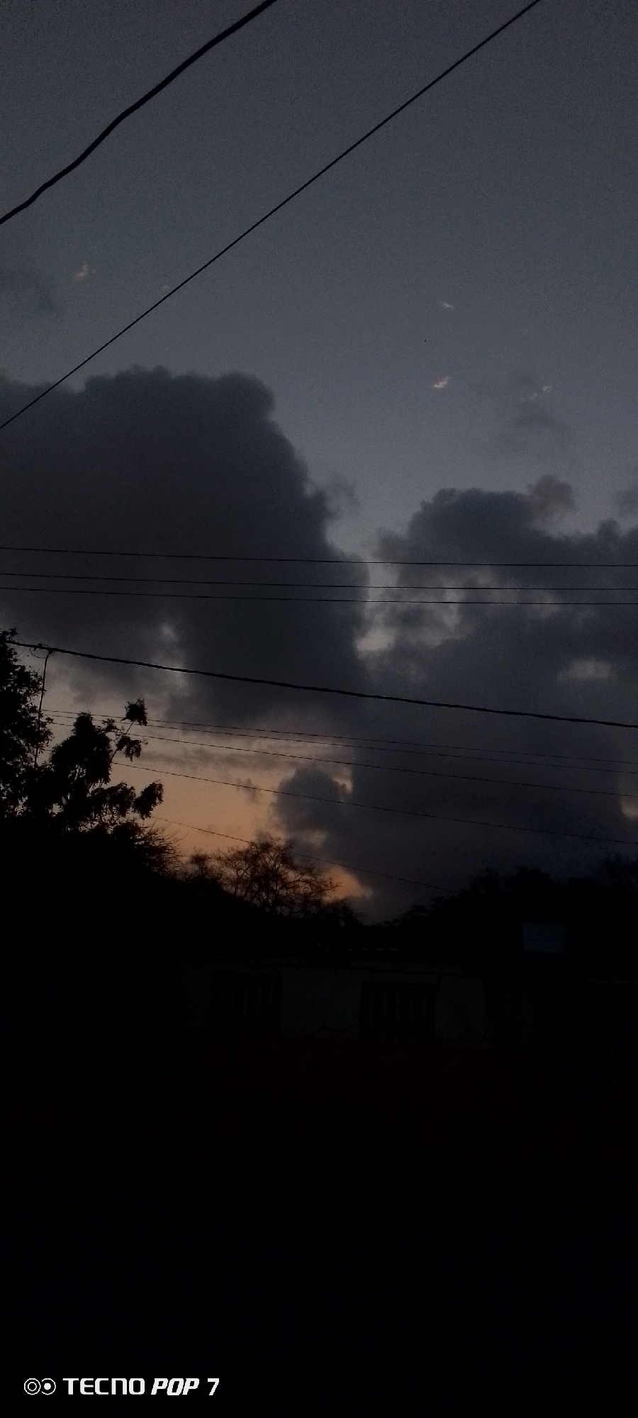 A low-angle shot captures a dark, overcast sky in Altagracia, Venezuela. Several electrical lines traverse the upper portion of the frame. Below, silhouetted trees and a building are visible against the fading light. A sliver of orange sky is visible between the clouds. The phrase "TECNO POP 7" is displayed in the lower-left corner.