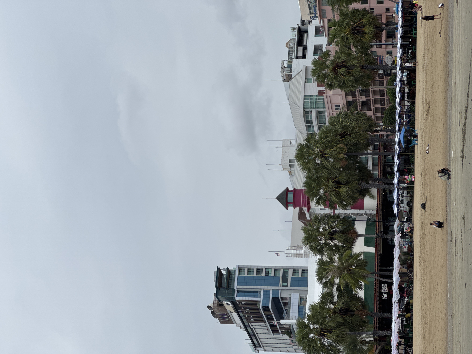 A cloudy day view shows a sandy beach in Pattaya City, Thailand, with the ocean not visible. In the foreground, several people are scattered across the sand, some sitting and others walking. Beyond the beach, a row of beach umbrellas and chairs is set up, with some people seated at tables beneath the umbrellas. Palm trees line the back of the beach, separating it from a line of buildings. The buildings vary in style and height, with a prominent modern high-rise with a "NIVIL" sign on top, a pink tower resembling a lighthouse, and other multi-story structures. The overall scene depicts a slightly overcast day at a popular beach resort.