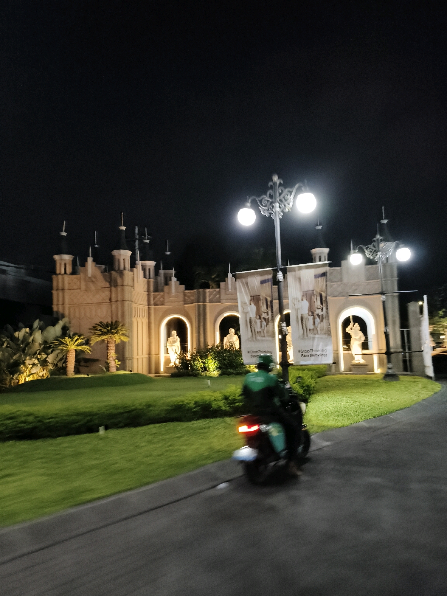 The image captures a nighttime scene outside a building that resembles a castle or a large ornate structure, likely located in Surabaya, Indonesia. In the foreground, a person is riding a motorcycle on a paved road. The motorcycle has its taillights illuminated. The building in the background is illuminated by artificial lights, highlighting its architectural features, including arches and statues. There are palm trees and manicured lawns in front of the building. Several large banners are hung between ornate lampposts in front of the building. The overall atmosphere is one of an illuminated, possibly festive, outdoor space at night.