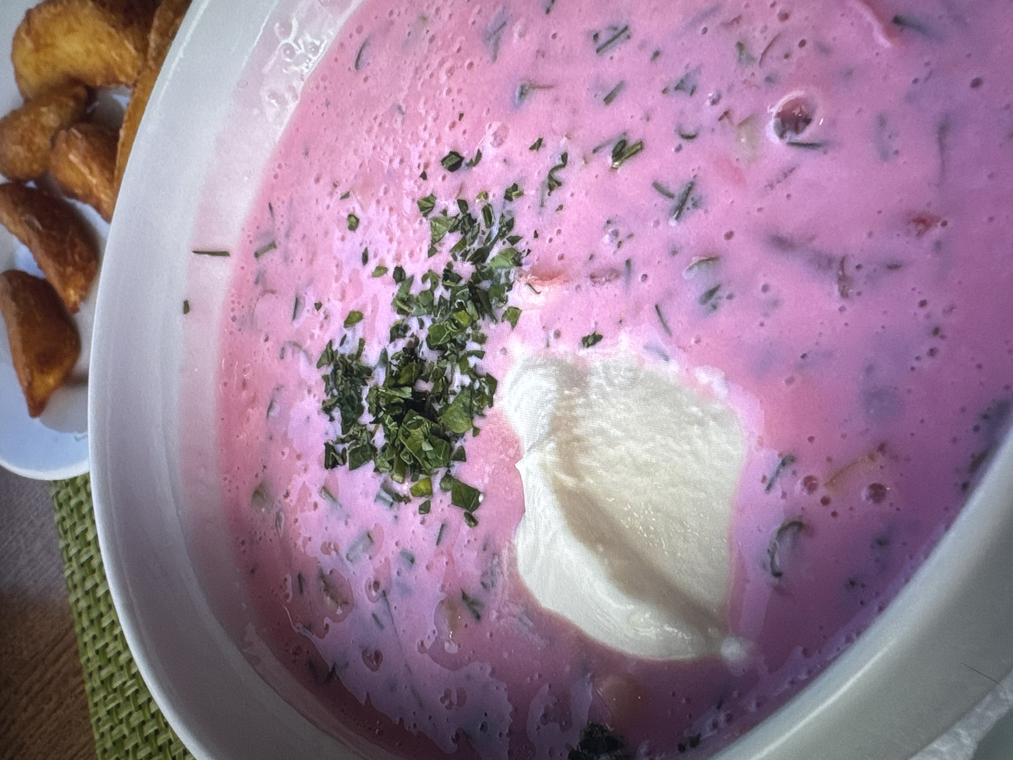 A close-up shot shows a bowl of cold beet soup, likely okroshka, with a dollop of sour cream and a sprinkle of fresh herbs. The soup has a vibrant pink hue, interspersed with finely chopped vegetables and herbs. In the background, a plate of golden-brown potato wedges is visible, suggesting a meal setting. The image does not provide information about the specific location in the city of Novinki, Russia.