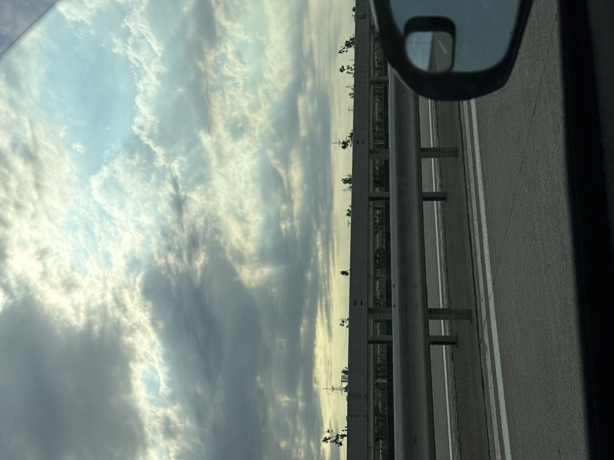 The image, taken in Surakhany Raion, Azerbaijan, captures a view from a vehicle, likely a car, traveling on a highway. The perspective is tilted, suggesting the photograph was taken with the camera held sideways.
The primary subjects are the sky and the roadside infrastructure. The sky is filled with dramatic clouds, a mix of bright, sunlit formations and darker, more ominous grey masses, hinting at a dynamic weather pattern. The sun appears to be low, possibly indicating either early morning or late afternoon, casting a soft, diffused light.
To the right of the frame, a grey highway barrier made of metal poles and horizontal beams runs parallel to the road. Beyond the barrier, a glimpse of distant structures and trees can be seen, suggesting an urban or semi-urban environment bordering the highway. A side-view mirror of the vehicle is visible in the upper right corner, reflecting a sliver of the road and sky ahead.
The overall mood is somewhat contemplative, with the vast, cloudy sky dominating the scene and the linear elements of the road and barrier providing a sense of movement and direction. There is no visible text in the image.