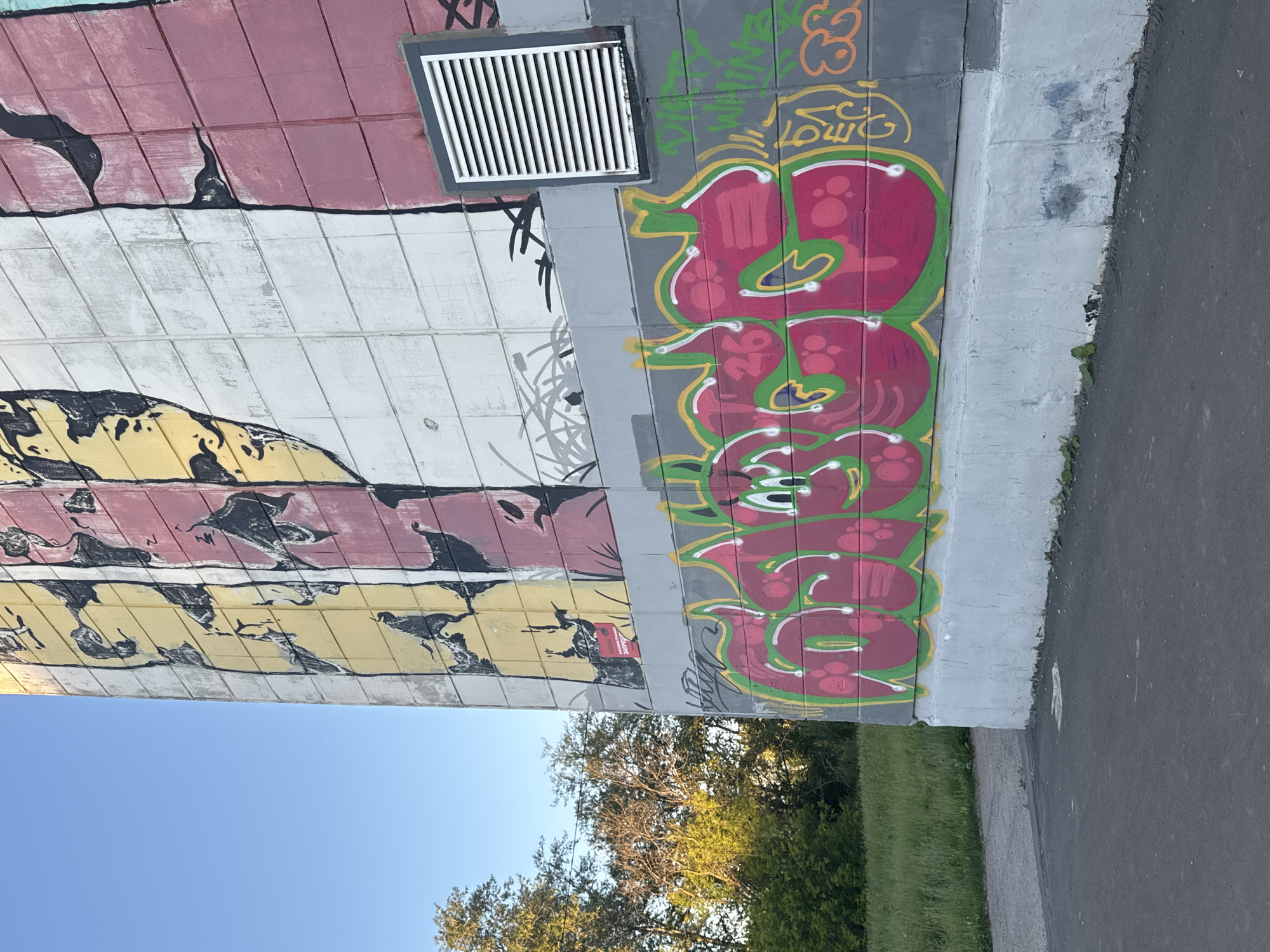 The image depicts a section of a building exterior featuring graffiti. The building appears to be a multi-story structure, possibly an apartment block, with a concrete tile facade. The graffiti is applied to the lower portion of the wall and covers a large, stylized lettering in bright pink and green. Above this, the wall is decorated with vertical stripes of yellow, pink, and white, with black abstract figures or patterns. A ventilation grate is visible on the right side of the building, mounted on the wall. The building is situated in an outdoor environment, with a grassy area and trees visible to the left. The sky is clear and blue. The overall scene suggests an urban setting, likely in a residential area. The graffiti itself is a prominent feature, dominating the lower part of the composition. The style of the graffiti is typical of street art, with bold lines and vibrant colors. The building's architecture, with its tiled facade, is characteristic of Soviet-era construction. The presence of trees and grass suggests a park or green space adjacent to the building. The image is captured in daylight, with natural lighting illuminating the scene. The perspective is from ground level, looking up at the building.