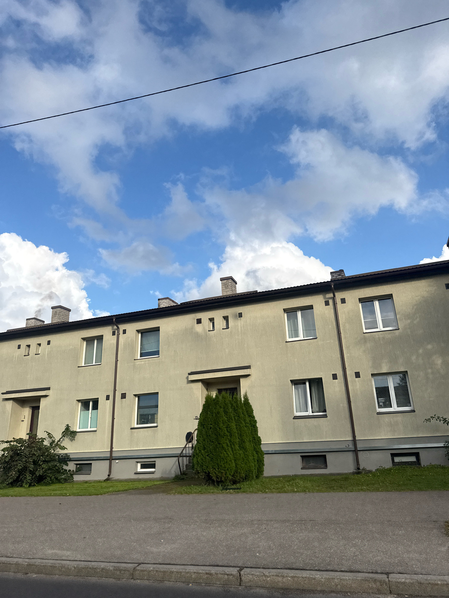 A light beige, two-story apartment building with a dark brown roof is shown in a suburban environment in Tallinn, Estonia. The building has multiple windows on both floors, most with white frames. There are also several small, square decorative elements and two evenly spaced chimneys on the roof. A dark gray asphalt road and sidewalk are in the foreground, with a patch of green grass separating the road from the building. Two mature evergreen trees flank the entrance of the building, one on the left and one on the right. A thin black wire extends diagonally across the top portion of the image against a blue sky filled with white, fluffy clouds. The scene is static and appears to be captured on a bright, clear day.