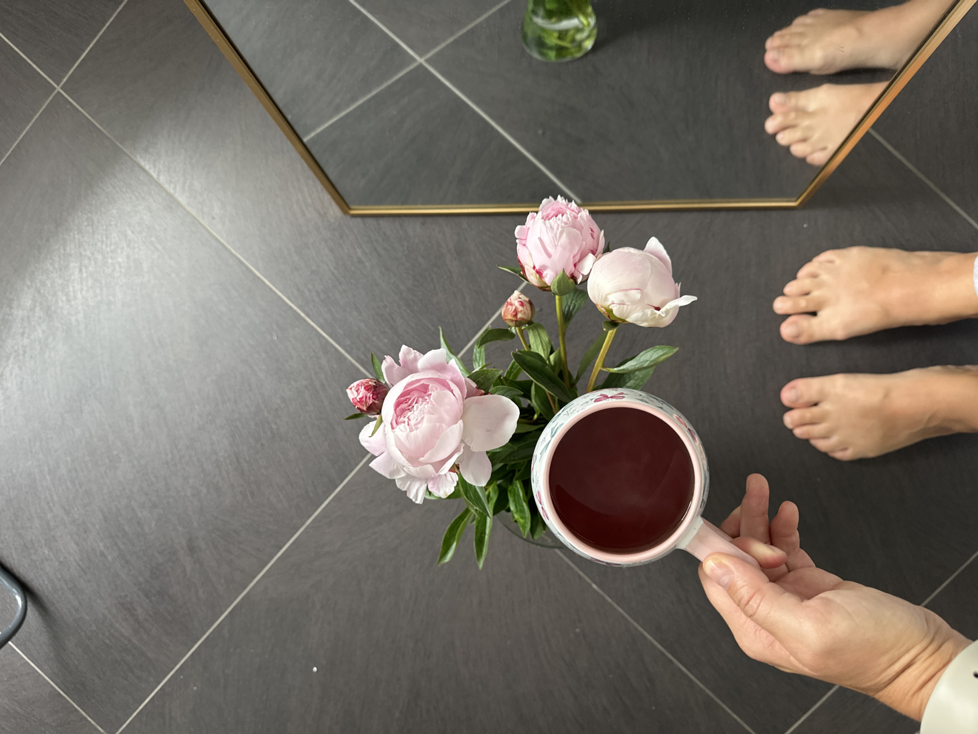 A top-down view of a person's feet on a dark tiled floor, next to a mug of dark liquid held in a person's hand. The mug is pink with a floral pattern. In the background, a bouquet of pink peonies sits in a vase. A gold-framed mirror reflects the feet, the floor, and a small green glass object. The scene is likely indoors, possibly in Kutenholz, Germany, given the context.