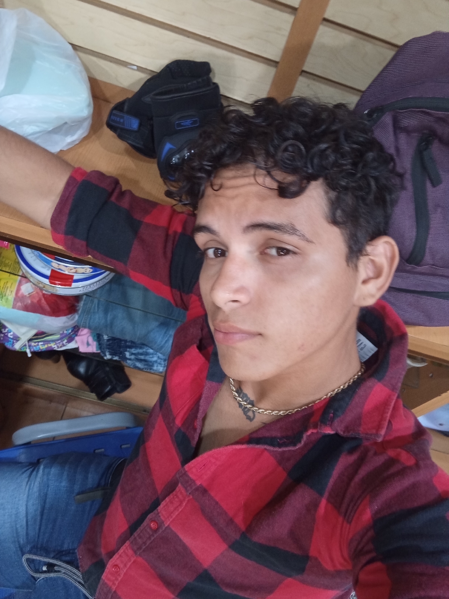 A young man with dark, curly hair and brown eyes is featured in a close-up selfie, looking directly at the camera with a neutral expression. He is wearing a red and black plaid button-up shirt, blue jeans, and a gold rope chain necklace. A dark tattoo is visible on his lower neck/upper chest, partially obscured by the necklace.
The setting appears to be indoors and somewhat cluttered. In the upper left background, a white plastic bag sits next to a pair of black gloves with blue accents. Above the man's head, to the right, a purple backpack is visible. Below his outstretched arm, various items are stacked, including what looks like a round metal tin with a colorful lid, folded blue jeans, and other clothing items. A black shoe or boot is also partially visible. The background consists of light-colored wooden panels, suggesting shelves or a wall. The man's body position implies he might be lying down or leaning back.
There is some faint text on the blue accents of the black gloves, possibly "TRICA" or a similar brand name, though it is not clearly legible. A white tag with black text is visible inside the man's shirt collar, but the text is unreadable. No other specific location cues or text are discernible from the image that would explicitly confirm the Juan Griego, Venezuela location. The lighting suggests an indoor daytime shot.