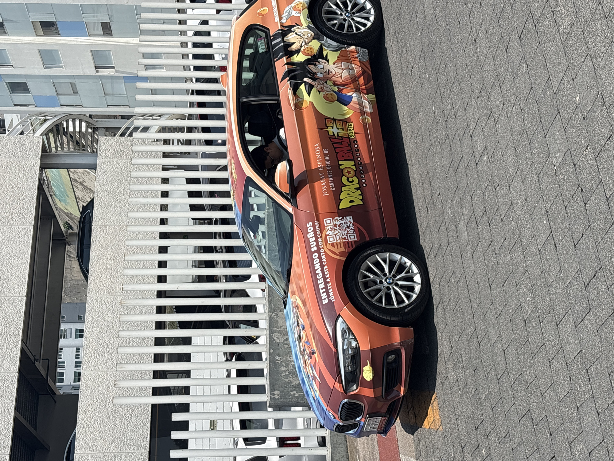 The image shows a car parked on a paved surface, likely a parking lot or a street, with a building in the background. The car is a BMW, and its side panels are decorated with a Dragon Ball Super theme, featuring characters and the logo of the popular anime series. Text on the car reads "JOSAFAT ESPINOSA CANTANTE OFICIAL DE DRAGON BALL SUPER" and "ENTREGANDO SUEÑOS ¡ÚNETE A ESTE CANTO CON CAUSA!". A QR code is also displayed on the car's side. A person is sitting inside the car. The license plate is partially visible. The background building is a modern structure with a white facade and a metal railing or fence. The car's license plate indicates it is from the State of Mexico, and the photo's location is in the city of Huixquilucan de Degollado, Mexico.