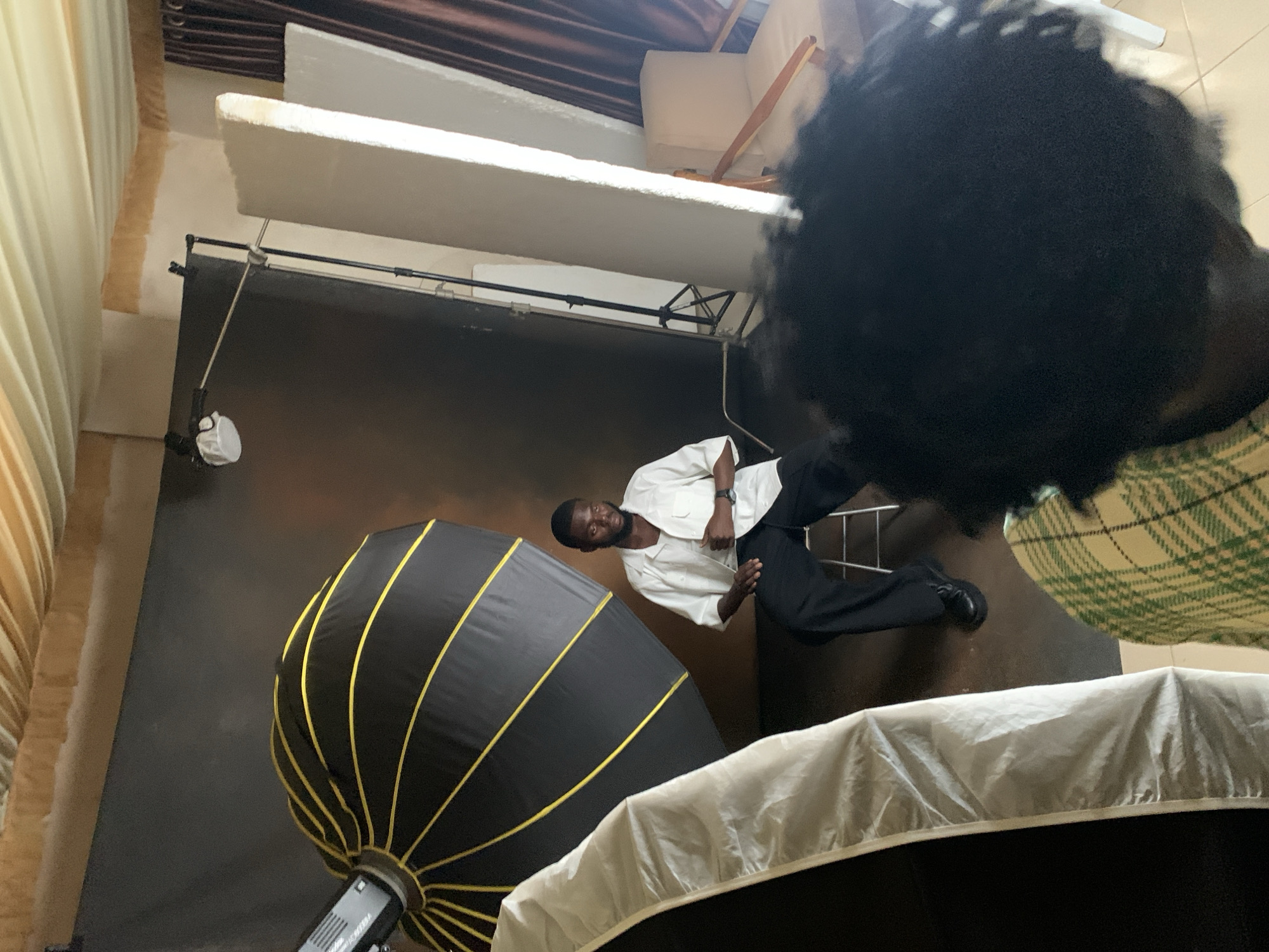 In a studio setting in Barakin Sabawa, Nigeria, a man is being photographed. He is dressed in a white collared shirt and dark pants, with his hands clasped in front of him. He is seated on the floor, facing towards the camera. To his left is a large, black octagonal softbox with yellow accents, a common piece of lighting equipment for photography. In the foreground, a white diffuser or softbox is visible, partially obscuring the bottom of the frame. The background is a dark, solid backdrop, typical for studio portraiture. Above the backdrop, a white panel hangs, possibly a reflector or a piece of the studio ceiling. The overall lighting suggests an indoor studio environment, likely during the day, as is typical for photo shoots. The man's expression is neutral, and the overall mood is professional. The scene is oriented vertically in the image, indicating the photograph was taken in portrait mode.