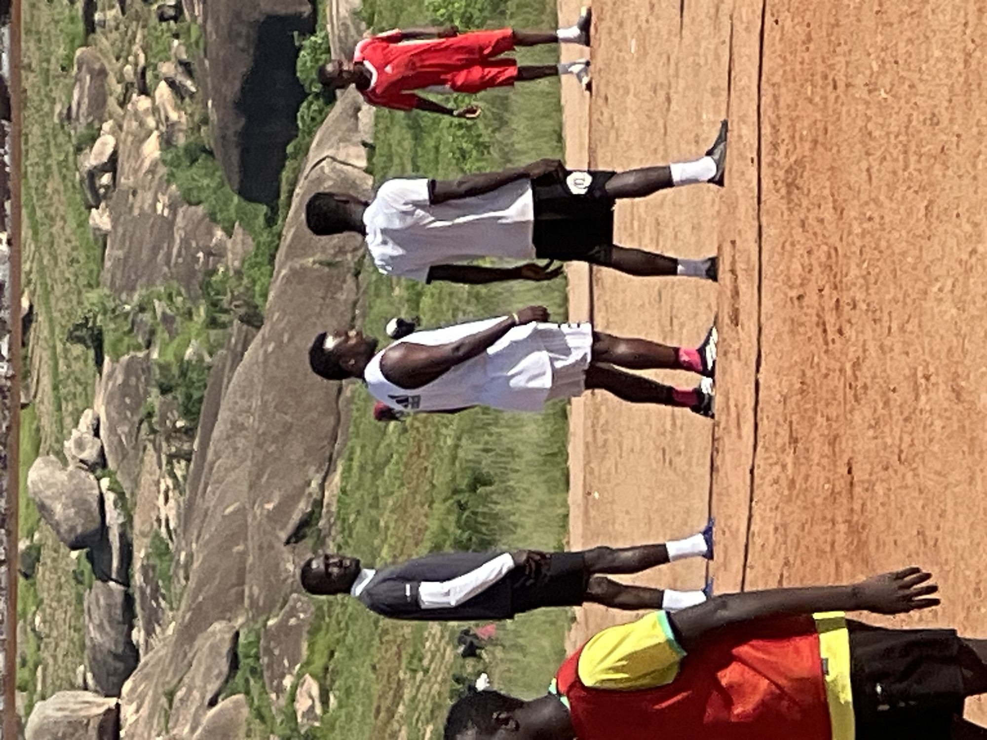 In the city of Jos, Nigeria, on a dusty, orange-colored field, four men are positioned. One man, on the left, wears a dark grey long-sleeved shirt with white trim on the collar and sleeves, black shorts, and white socks. He is looking to his left. To his right, a man in a white t-shirt and white shorts, with a logo on his left chest, is facing away from the camera. Next to him, a man in a white t-shirt and black shorts with a white logo is also facing away. Further to the right, a man in a red t-shirt and red shorts, with white socks and black shoes, faces the camera. In the foreground, on the far left, partially visible, is a person wearing a red and yellow vest over black shorts. The background features rocky terrain with scattered green vegetation.
