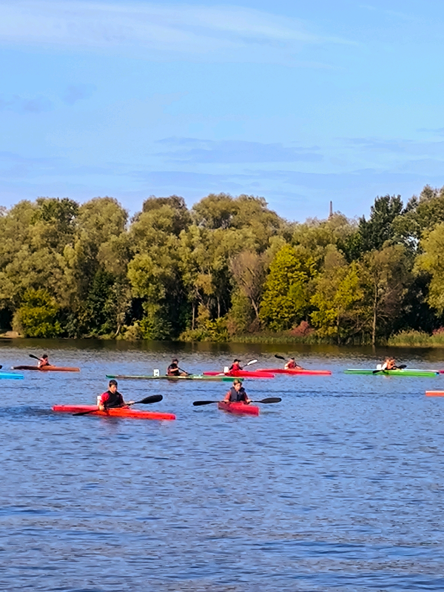 A group of kayakers is seen on a body of water, with a lush, tree-lined bank in the background. The sky above is a clear, pale blue with a few wispy clouds. In the distance, a tall, narrow structure, possibly a monument or spire, is visible above the trees. The kayakers are paddling in various colored kayaks, including red, blue, and green. Some of the kayakers are wearing life vests. The water is calm and reflects the colors of the sky and the surrounding foliage. The overall scene suggests an outdoor recreational activity taking place in a natural setting within or near a city.