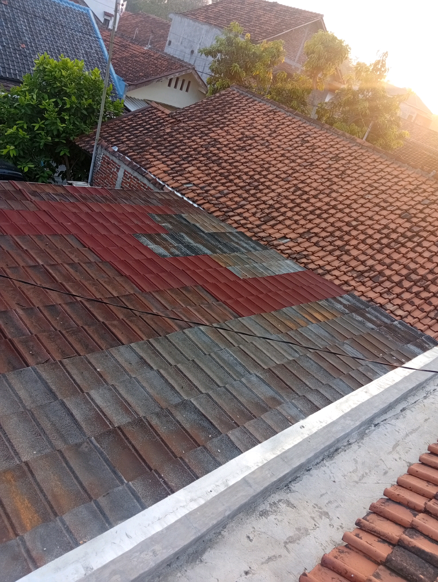 The image shows a rooftop view of residential buildings in the city of Kebandingan, Indonesia. The foreground features a section of roof with varying colored tiles, some appearing weathered and possibly aged, with a light-colored concrete edge along the bottom. Behind this, a larger area of traditional orange clay roof tiles is visible, leading to more rooftops and trees in the background. The setting appears to be during daytime, with sunlight casting a warm glow and lens flare in the upper right portion of the image. The overall scene depicts a typical urban or suburban residential environment.