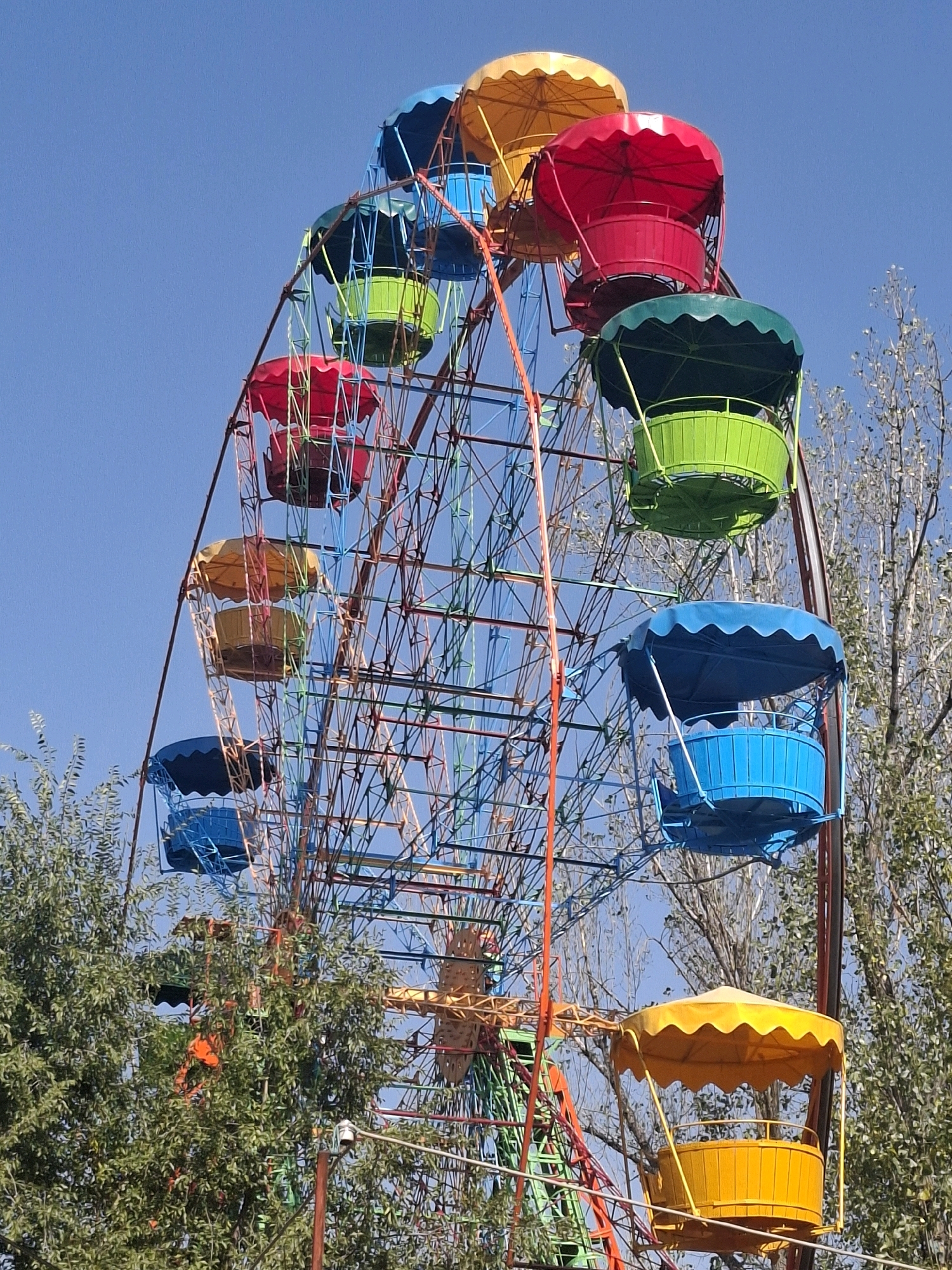 A brightly colored Ferris wheel dominates the frame against a clear blue sky. The Ferris wheel has multiple gondolas, each a different vibrant color including yellow, red, green, and blue. The structure of the Ferris wheel is a complex network of metal bars painted in various shades of red, blue, green, and yellow. Trees with green foliage are visible on the left and right sides of the frame, partially obscuring the lower parts of the Ferris wheel. In the background, the bare branches of deciduous trees are visible against the sky. The lighting suggests it is daytime, likely outdoors in a park or amusement area. The scene appears to be located in Almaty, Kazakhstan, likely within a public recreational space.