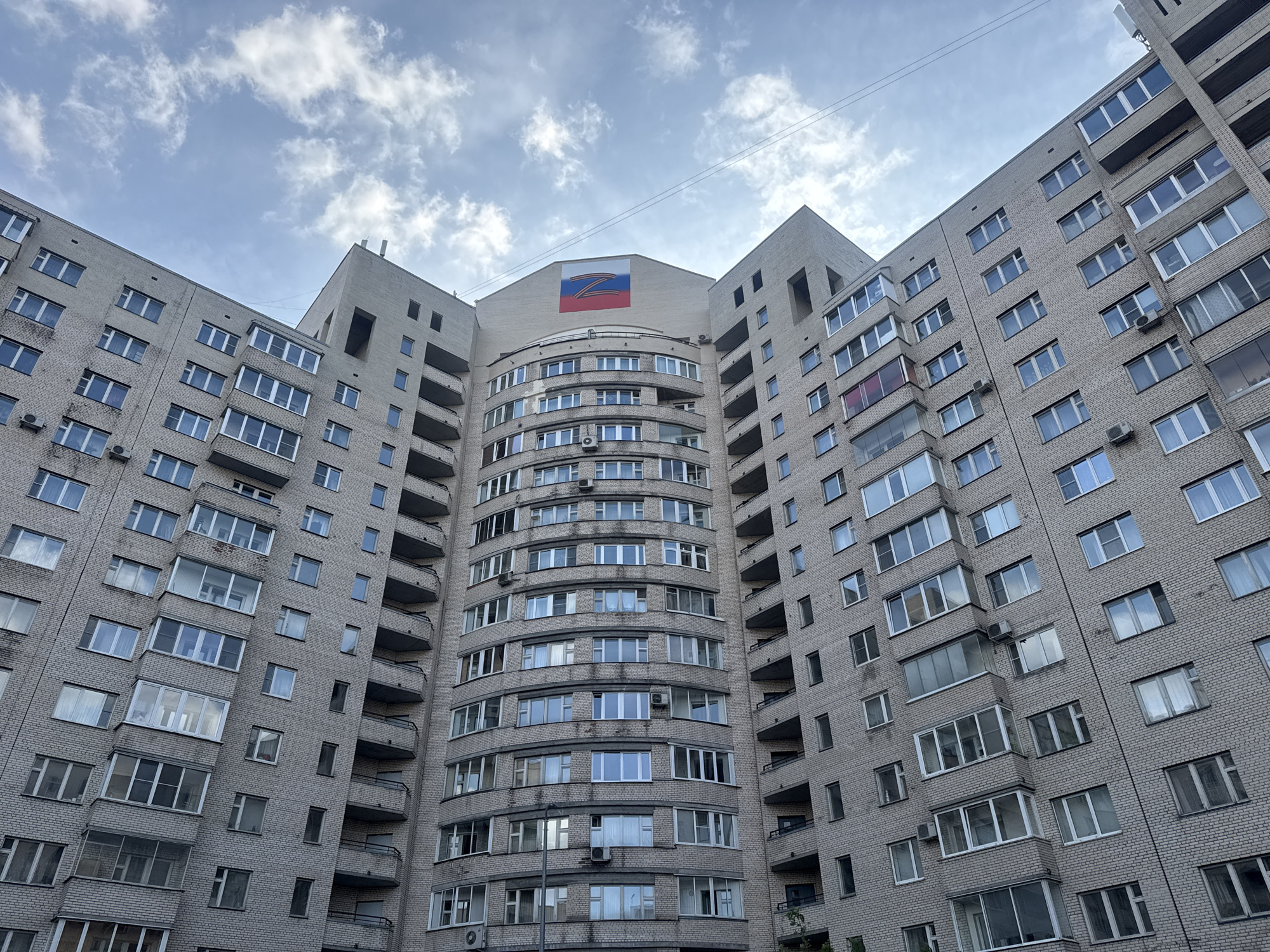 The image shows a tall, multi-story apartment building in Saint Petersburg, Russia, constructed of light gray brick. The building has a symmetrical design, with two wings flanking a rounded central section. Many windows, some with balconies, are visible on each floor. At the top of the central section is a large rectangular symbol featuring the Russian flag colors (white, blue, red) with a black and orange ribbon and a stylized "Z" on it. The sky is overcast with scattered clouds.