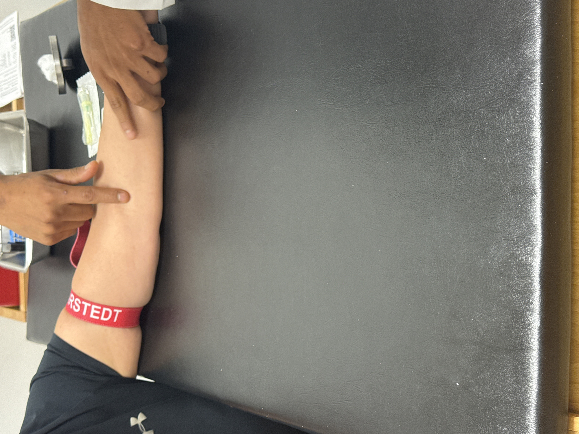 The image captures a close-up of a medical procedure, likely blood drawing or IV preparation. A patient's arm, clad in a black athletic sleeve, rests on a black, textured examination table or medical bench. A red tourniquet, with the partial text "RSTEDT" visible, is tightly wrapped around the patient's upper arm.
Two hands, presumably belonging to a healthcare professional, are seen interacting with the patient's forearm. One hand is palpating the area of the antecubital fossa, while the other is positioned higher up the arm, seemingly stabilizing it. On the table next to the arm, various medical supplies are visible, including a sterile-packaged needle or cannula, and a crumpled white gauze or tissue. A small metallic container is partially visible in the upper left corner of the frame.
The setting appears to be a medical or clinical environment. The lighting is neutral, providing no specific time-of-day cues. The context provided for this image is San Bernardino Tlaxcalancingo, Mexico.