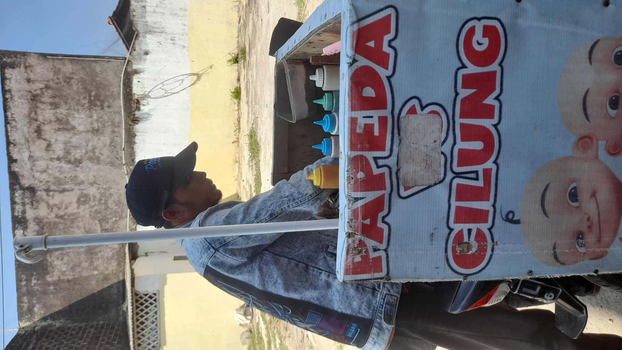 An Indonesian man wearing a denim jacket and a black baseball cap stands behind a food cart in Selaawi, Indonesia. The cart is adorned with red lettering that spells "PAPEDA CILUNG" and features images of cartoon babies. Several condiment bottles, including yellow, blue, green, and white, are visible within the cart. The background shows a weathered building and a clear blue sky. The man appears to be preparing food for customers, though no customers are visible in the frame.