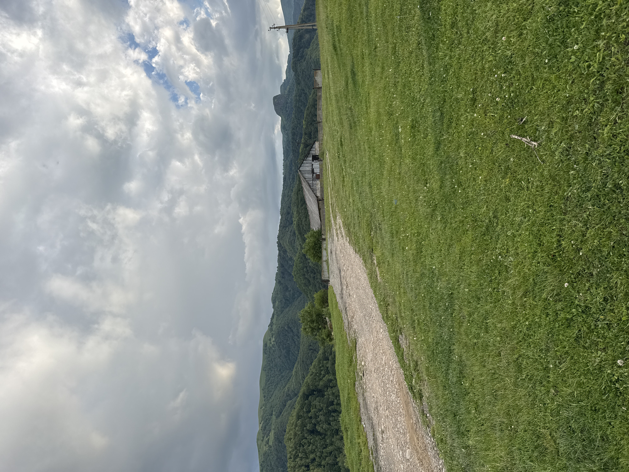 The image depicts a rural, hilly landscape under a cloudy sky. In the foreground, a dirt road curves from the left towards the center of the frame, bordered by a grassy field. In the middle ground, a long, rustic wooden building with a sloped roof stands to the right of the road, with a low fence in front of it. Beyond the building, a distinct, rocky peak rises from the densely forested mountains that dominate the background. The sky is filled with dramatic, gray clouds, with some lighter patches suggesting breaks in the overcast. A wooden utility pole with wires is visible on the far right. The scene is devoid of any people or animals. The environment appears to be a natural, undeveloped area.