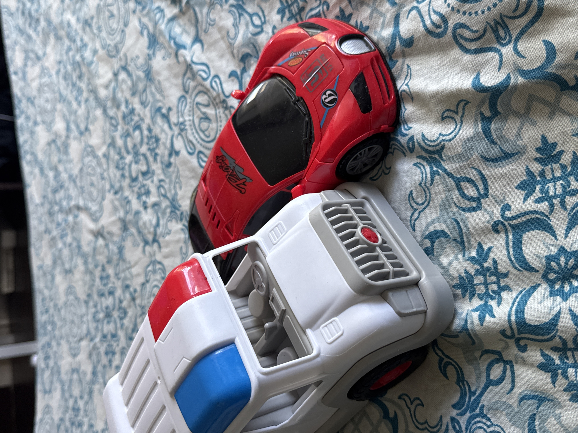 Two toy vehicles, a white ambulance-style truck with blue and red lights on top and a red sports car, are positioned on a patterned blue and white fabric surface. The fabric has a damask-like design with floral and swirling motifs. The ambulance toy is on the left, its front grille visible, and the red sports car is on the right, angled towards the viewer. The image is a close-up shot, and the background is blurred. The location of Яромінскі сельскі Савет, Belarus is not depicted in this image, as it is a still life of toys.