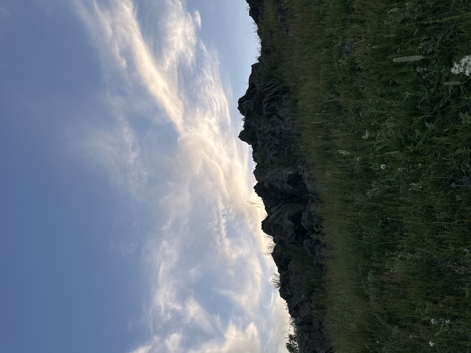 The image depicts a side view of a rocky, grassy incline against a backdrop of a sky filled with wispy clouds. The rock face is dark and jagged, appearing to be part of a cliff or hillside. Green grass and small white flowers grow along the top of the incline and spill down its sides. The sky is a soft blue with streaks of white clouds that are illuminated by a warm light, suggesting either sunrise or sunset. The overall mood is serene and natural. There is no text visible in the image.