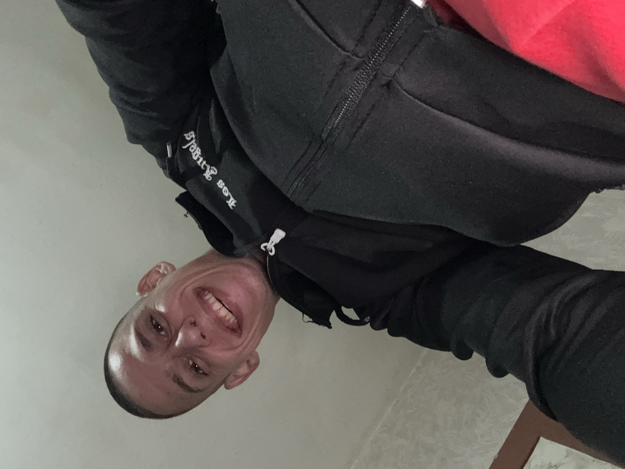 A man with a shaved head is smiling at the camera. He is wearing a black zip-up hooded sweatshirt with "Los Angeles" written on the sleeve. The background is a plain, light-colored wall and a tiled floor. There is not enough information to determine a location in Sofia, Bulgaria.