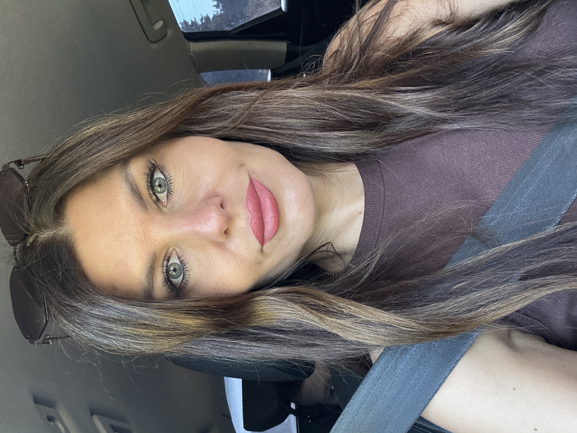 The image is a close-up selfie taken by a woman with long, brown hair with highlights, sitting in the driver's seat of a car. She is wearing a brown tank top and sunglasses are perched on her head. The seatbelt is visible across her chest. Her eyes are open and she is looking at the camera with a neutral expression. Her makeup includes eyeshadow, mascara, and lipstick. A subtle nose piercing is visible. In the background, a child can be seen in the back seat. The overall environment appears to be a car interior. No specific location within Noviy, Russia is depicted.