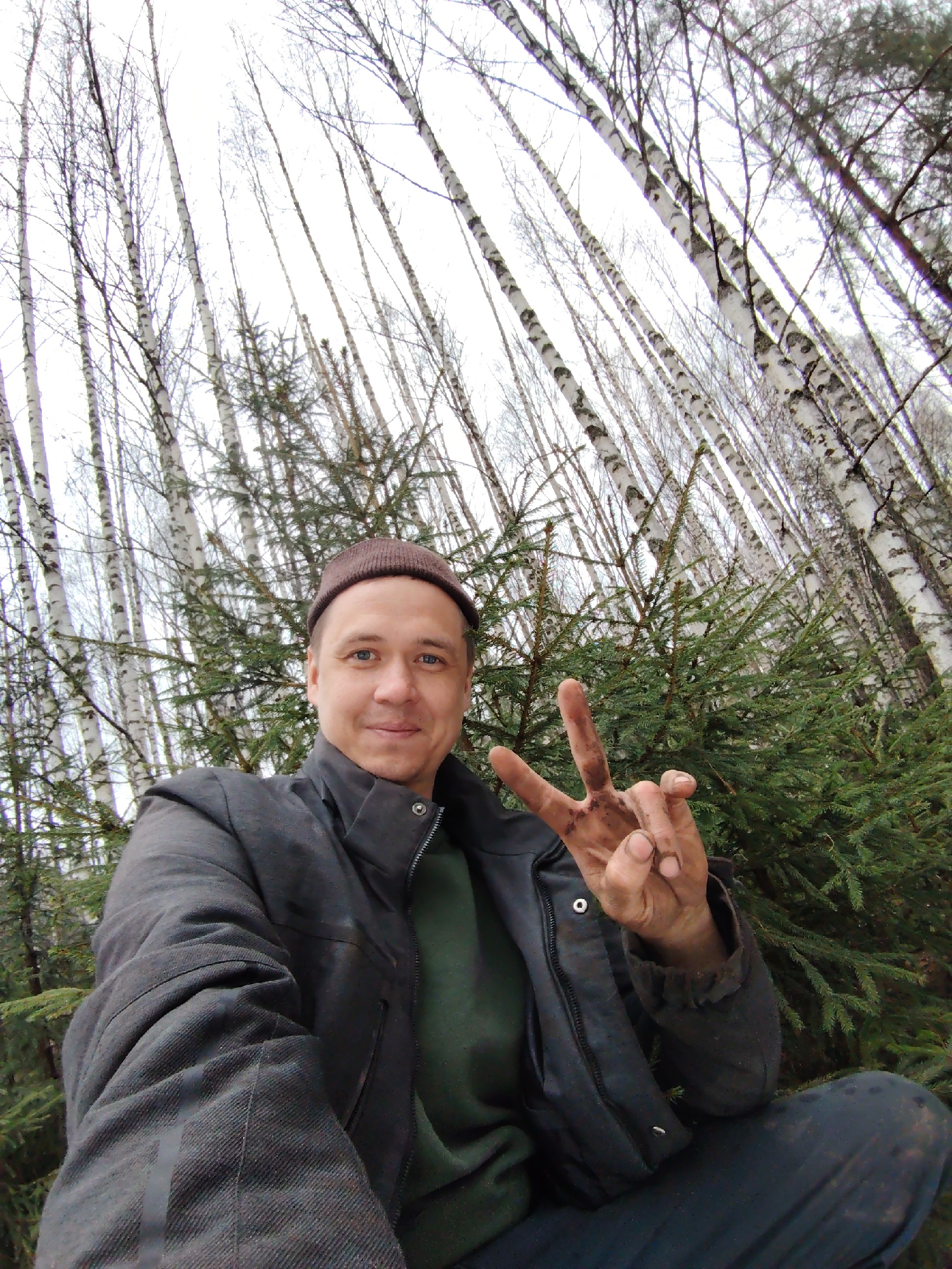 This is a selfie of a man taken outdoors in a forest, identified as Markovskii, Russia. The man, appearing to be in his 30s or 40s, has short dark hair and blue eyes, and is wearing a dark brown knit cap, a dark grey or black jacket over a dark green sweater. He is smiling faintly at the camera while making a peace sign with his right hand. A notable detail is that his hand is visibly dirty with what appears to be soil or mud.
The background consists of a dense forest. Numerous tall, slender birch trees with white and black bark stand prominently, their branches mostly bare, suggesting a time of year like late autumn, winter, or early spring. Below and among the birches are several smaller, green evergreen trees, likely fir or spruce. The sky is bright and overcast, providing diffused and even lighting across the scene. The overall atmosphere conveys a quiet, natural setting. There is no visible text in the image.