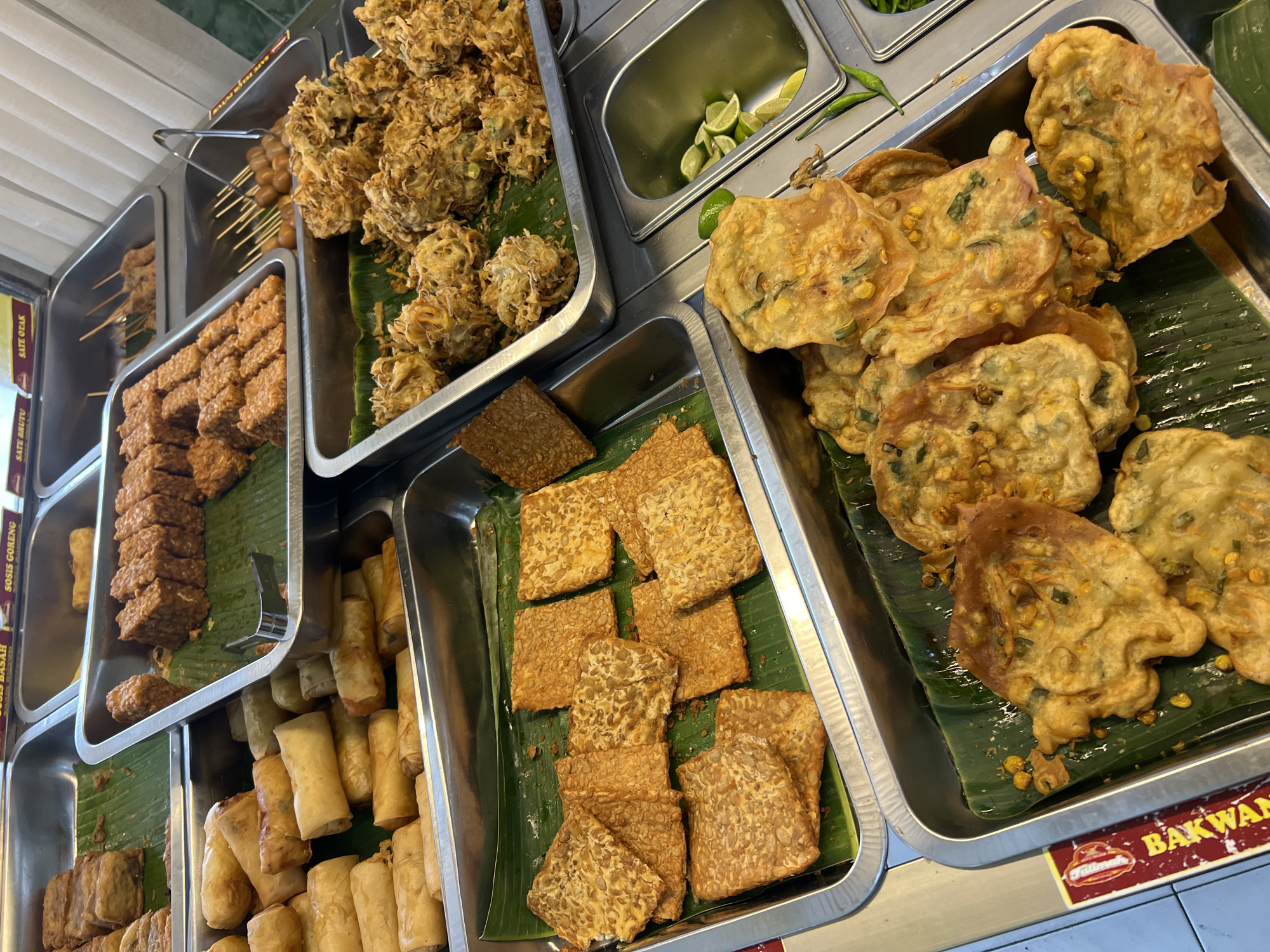 An overhead shot captures a vibrant display of Indonesian street food at a food stall located in the city of Condongcatur, Indonesia. The scene is dominated by various fried items arranged in rectangular metal trays lined with banana leaves. In the foreground, a tray is filled with irregularly shaped, golden-brown fried fritters, possibly bakwan, some of which are partially obscured by others. These fritters appear to contain visible pieces of corn and green vegetables. Adjacent to this tray is another, slightly elevated one, containing a collection of perfectly square, sliced pieces of tempeh, a fermented soybean product, also fried and golden. Further back, to the left, a tray holds numerous golden-brown spring rolls, some whole and some cut in half. To the right of the tempeh tray, another tray displays a mound of crispy, irregular fried morsels, resembling vegetable fritters or tempura. Above these, a tray contains skewers of small, round items, possibly meatballs or fish balls, and another tray shows rows of rectangular fried items, perhaps fried tofu or a similar dish. The background is blurred, but suggests a bustling food stall environment with other food offerings visible. The lighting is bright and even, highlighting the textures and colors of the food. There are no people visible in the frame.