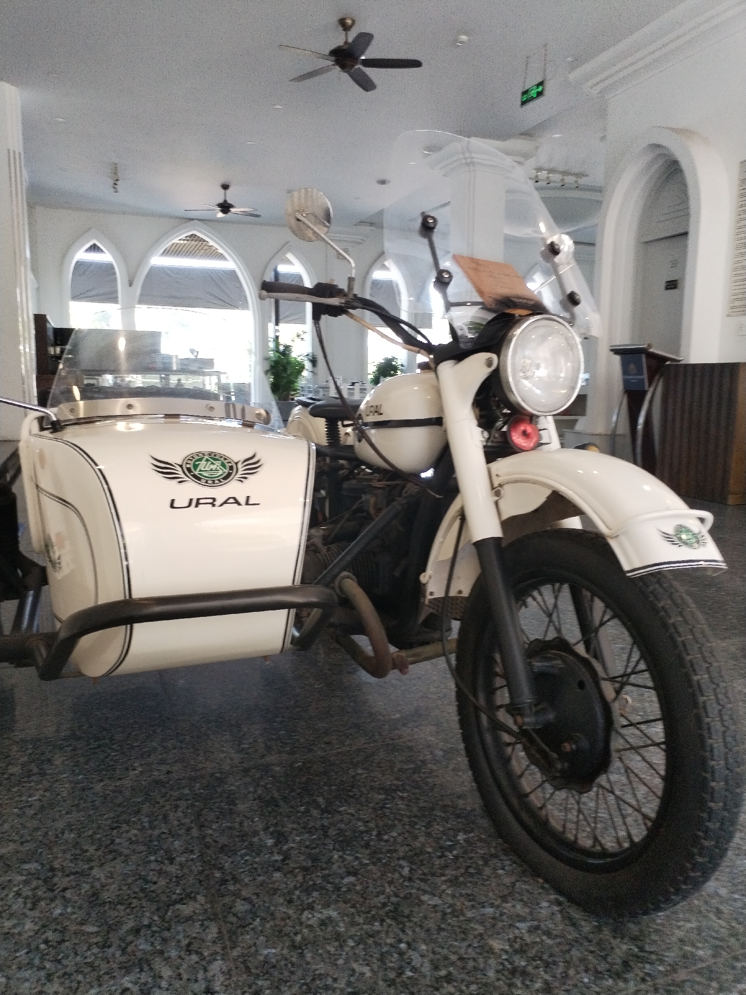 The image shows a white Ural motorcycle with a sidecar parked indoors, on a speckled grey floor. The motorcycle is positioned in the foreground, with its front wheel and headlight prominently displayed. The sidecar is attached to the left side of the motorcycle and has the word "URAL" written on it, along with a logo. The background reveals an indoor space with arched windows and ceiling fans. The architecture suggests a public building, possibly a hall or a restaurant, with white pillars and high ceilings. There is a green exit sign visible in the upper right portion of the image. No people are visible in the scene. The overall lighting is bright, indicating daylight, possibly filtered through the windows. The location is identified as Bac Nha Trang Ward, Vietnam.