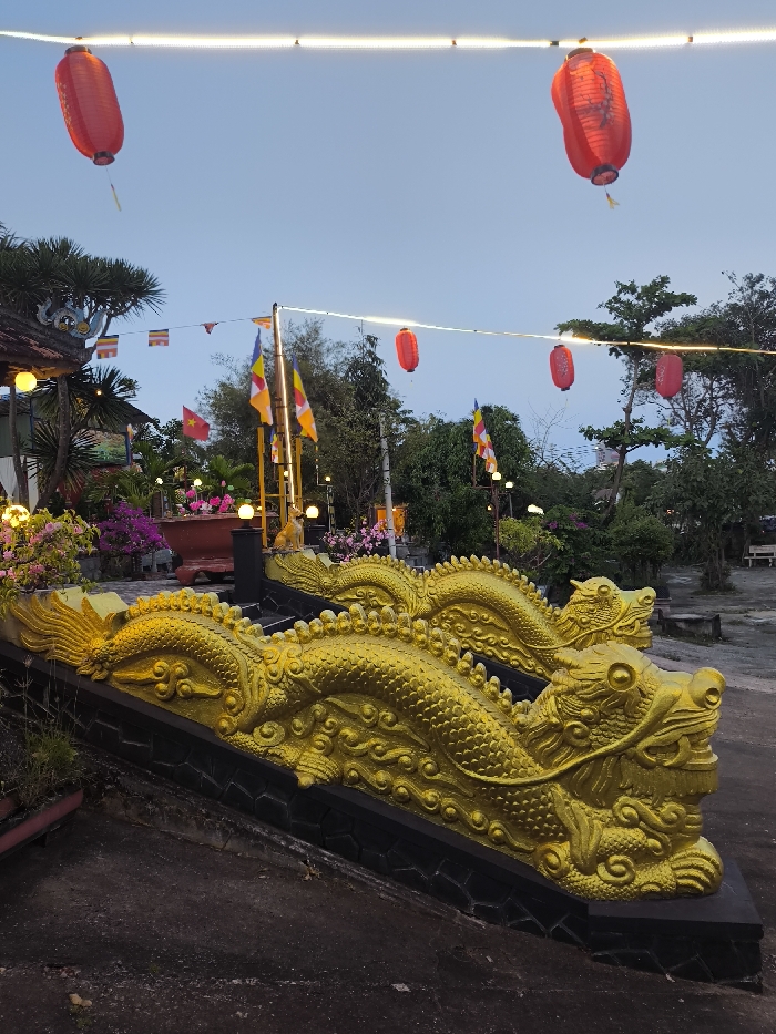 The image depicts an outdoor scene at twilight within Tuy Hòa, Vietnam, focusing on an ascending pathway flanked by ornate golden dragon balustrades. Two prominent, intricately scaled golden dragon sculptures with visible claws form the balustrade on a black, textured base, terminating in detailed dragon heads facing right in the foreground. The ground immediately below is dark, worn pavement. Overhead, multiple string lights and five cylindrical red lanterns are suspended across the frame, two of which display dark script.
Behind the balustrades, a landscaped garden area is visible, featuring abundant pink and purple flowering plants, consistent with bougainvillea, interspersed with green foliage. Several glowing spherical lights are embedded within the garden. A large reddish-brown planter and a small, light-colored, possibly canine-shaped statue on a dark pedestal are also present. A tall pole displays multiple Buddhist flags (vertical stripes of yellow, white, orange, blue, and red) and a single red Vietnamese flag with a yellow star. Further red lanterns and string lights are scattered throughout this midground. The background consists of dense green trees and brush, with indistinct structures partially visible through the foliage on the right. A simple wooden bench is situated under trees on the far right. The sky is a pale blue-grey, indicative of dusk. No individuals are present.