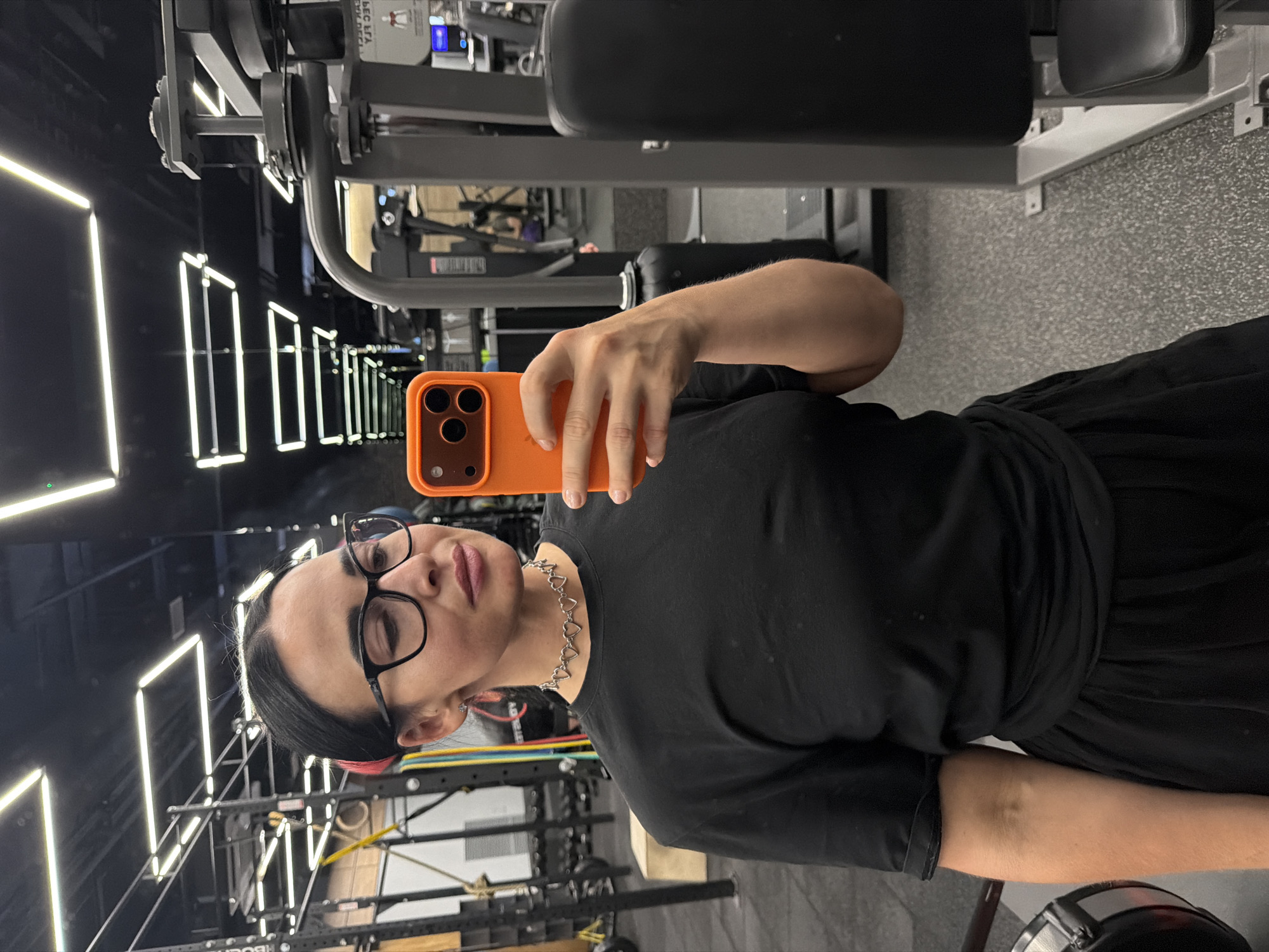A person with black hair and glasses is taking a selfie with an orange phone in a gym. The person is wearing a black t-shirt and a heart-shaped necklace. In the background, there is gym equipment and illuminated light fixtures. The location is not discernable from this image.