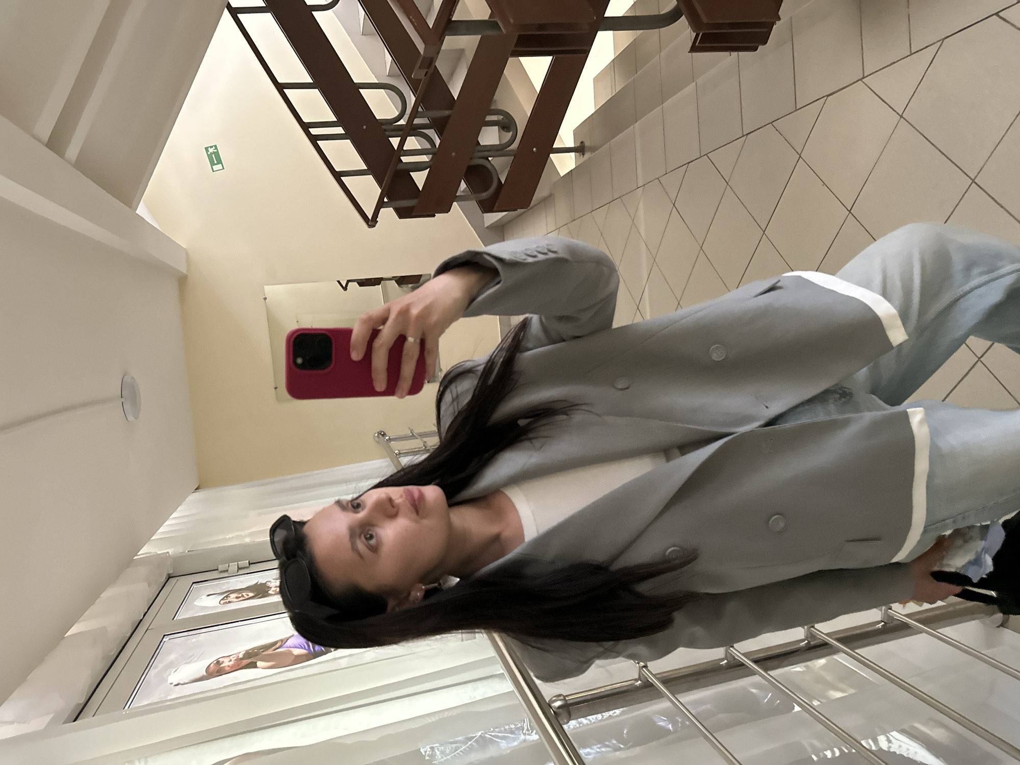 A woman with long, dark hair, wearing a light grey blazer, white top, and light-wash jeans, is taking a selfie with a red smartphone. She is standing in front of what appears to be a display window or a series of framed posters depicting other women. Behind her are stairs with a brown metal railing, and a tiled floor suggests an indoor setting, possibly a public building in Chaikovsky, Russia. An exit sign is visible on the wall near the stairs.
