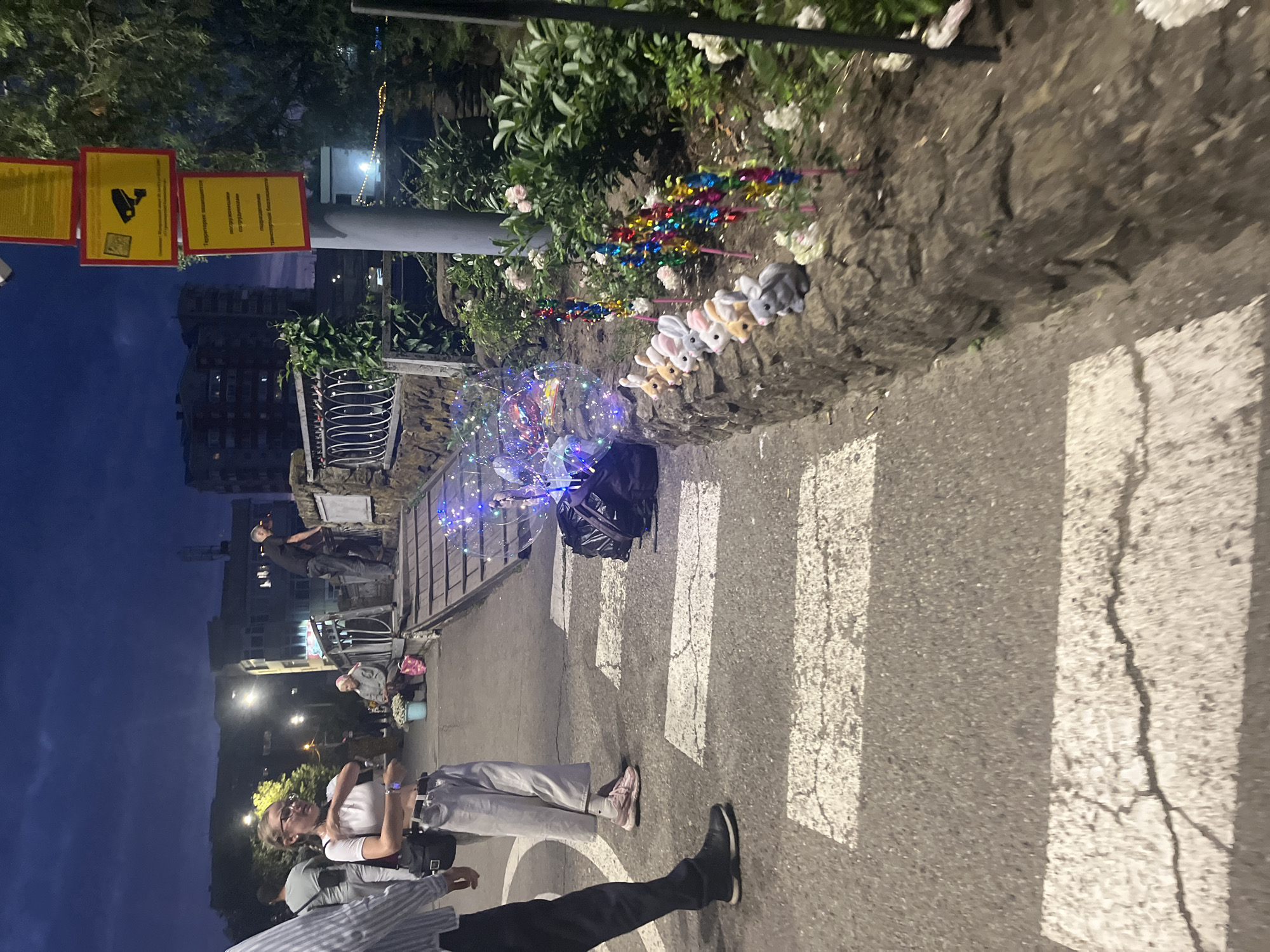 This image depicts a street scene at dusk in Krasnodar, Russia. The foreground shows a pedestrian crossing with white stripes on asphalt. To the right of the crossing, a low stone wall borders a garden area where several plush rabbit toys and colorful pinwheels are displayed. A person is walking across the street, while another, a young woman with glasses wearing a white crop top and light-colored pants, stands near the crossing, looking to her left and gesturing with her hands. In the background, there are buildings, streetlights, and trees. Several signs are visible on a pole to the right, one featuring a security camera icon and text in Russian. The overall atmosphere is that of a public urban space during twilight hours.