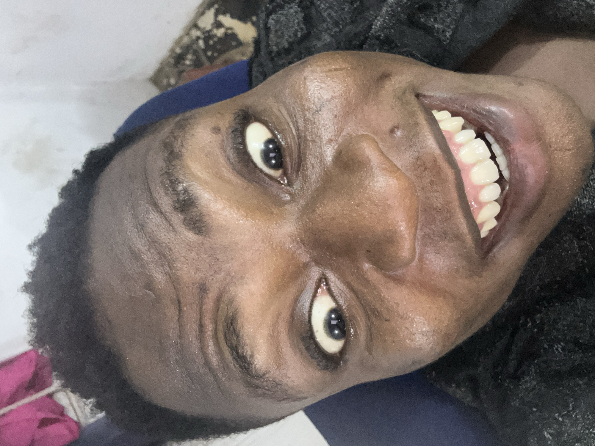 A close-up, rotated image shows a person with dark skin and short, dark hair smiling widely. The person's teeth are visible. The person is wearing a dark-colored, textured shirt. The background is a mix of lighter colors, suggesting a wall, and a dark blue area, possibly a chair. A partially visible pink object is present in the lower left corner. The location and city are Unknown, Unknown.