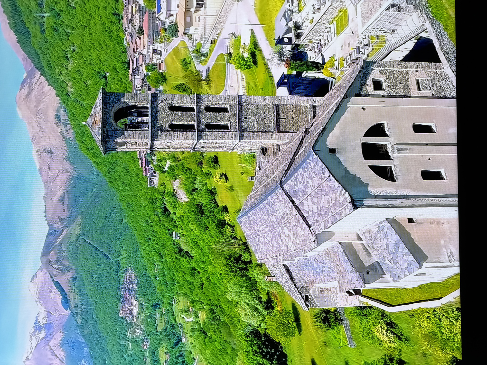 This is an aerial, eye-level view of a stone church with a bell tower in a mountainous, green valley. The church has a gray, slate roof and a light-colored stone and stucco facade. It features arched windows and smaller rectangular windows. The bell tower is square, made of stone, with arched openings at the top and rectangular openings on lower levels. In the background, a village with red-tiled roofs is nestled on a hillside, with more houses scattered across the green, forested slopes. Snow-capped mountains form the distant backdrop. The immediate foreground shows a cemetery adjacent to the church. The scene is brightly lit with sunlight. The image provided does not show the city of Chirchik, Uzbekistan.