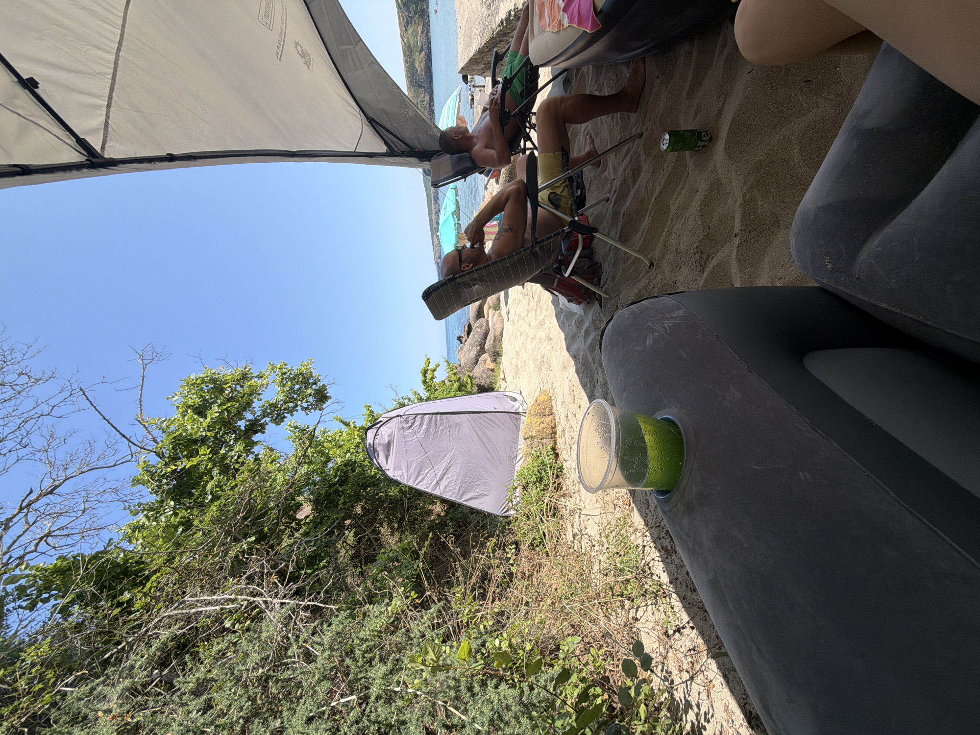 The image captures a sunny day on a beach in Sinemorets, Bulgaria. In the foreground, a large, dark gray inflatable couch with a cup holder containing a green drink is visible. To the left, dense green foliage and dry bushes grow on a sandy embankment. Further back, a gray portable changing tent stands near the vegetation. Two men are relaxing in beach chairs under a light-colored canopy, with one man wearing yellow shorts and the other wearing green shorts. The beach sand extends towards the sea, where the calm blue water meets a distant, hilly coastline. The sky is clear and bright blue. A green can of Heineken beer is on the sand near the chairs.
