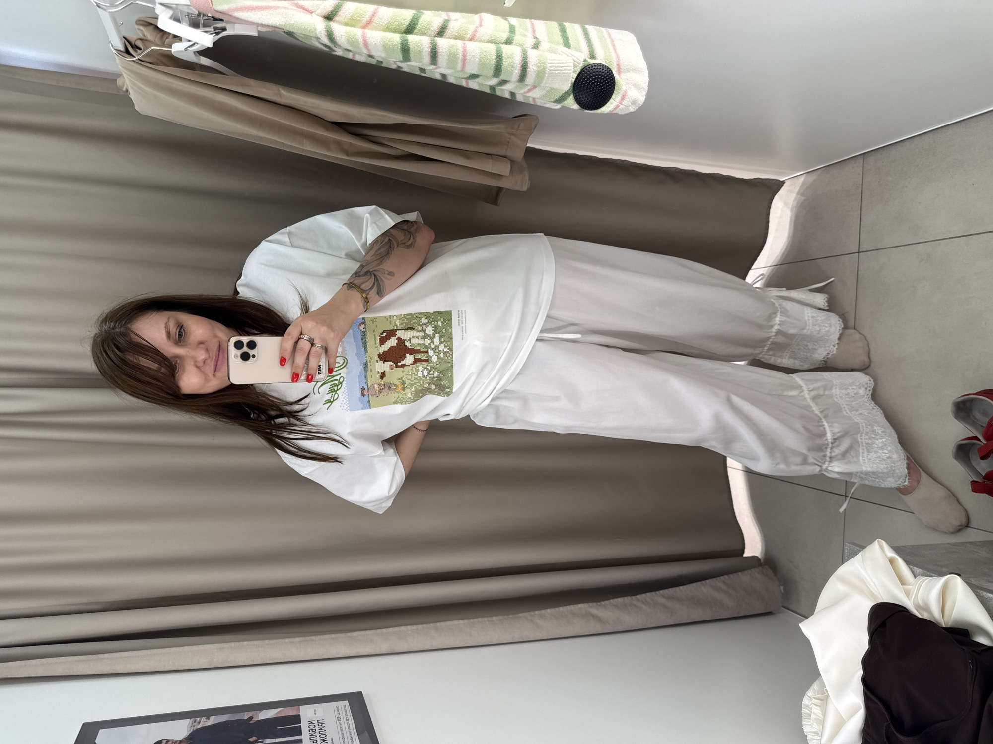 A woman is standing in a fitting room, holding a smartphone and taking a selfie. She is wearing a white t-shirt with a graphic print of a cow in a field, and white pajama-style pants with lace trim. In the background, there are beige curtains, clothes hanging on a rack, and a poster on the wall. The floor is tiled. The scene appears to be in a retail store or a fitting room. The image has no discernible connection to the city of Sochi, Russia.