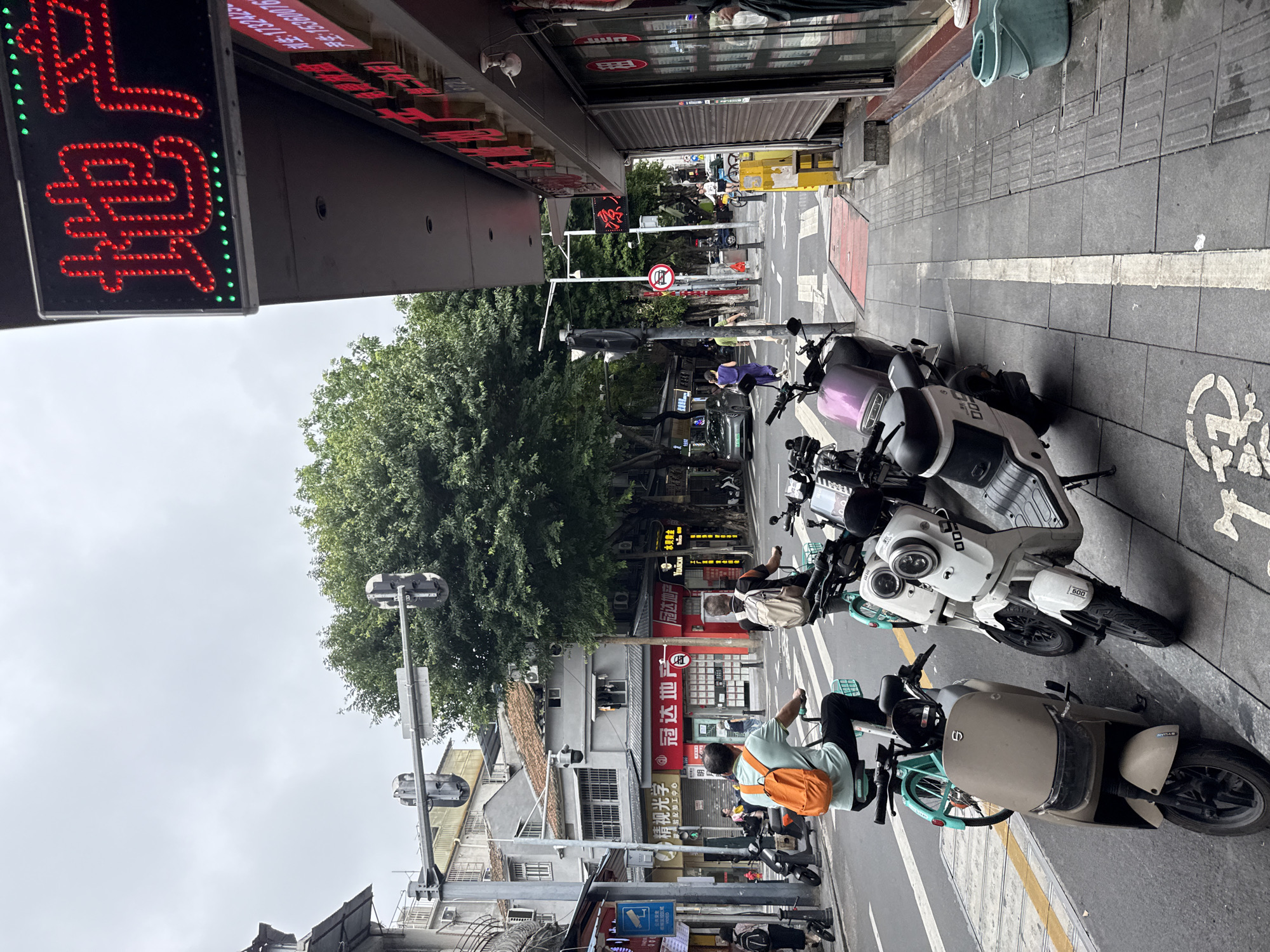 The image shows a street scene in Liwan District, China, on an overcast day. In the foreground, several parked electric scooters and a bicycle are visible on the sidewalk. A man wearing a light blue shirt and an orange backpack is riding a bicycle in the street. Another person, also wearing a backpack, is riding a scooter behind him. The street is paved with asphalt and has painted lane markings. On the right side of the street, there is a building with a red awning and a neon sign displaying Chinese characters. The sign is illuminated with red and green lights and appears to be advertising real estate. Shops and other buildings line the street, and trees with lush green foliage are scattered throughout the scene. Traffic lights and street signs are also visible. The overall impression is of a bustling urban environment with a mix of traditional and modern elements.