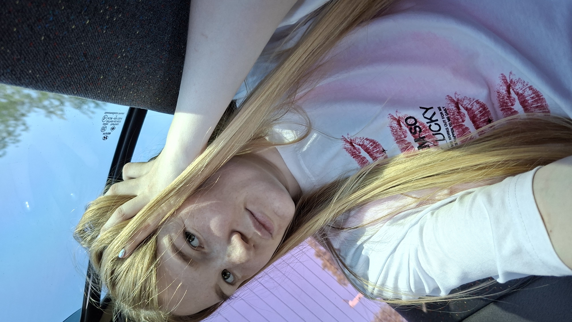 A young woman with long blonde hair is angled in a photo taken inside a vehicle, seemingly from a passenger seat perspective. She rests her head on her hand, her face angled towards the camera. She is wearing a white t-shirt with red lipstick kiss prints and the words "LUCKY MSO". The vehicle's interior includes a dark-colored ceiling and a blue tinted window reflecting the sky and some trees. Part of the vehicle’s frame and potentially another surface (perhaps a car seat or dashboard element) are visible in the foreground with a purplish reflection. The location in the city of Большой Букор, Russia, cannot be verified through the image.