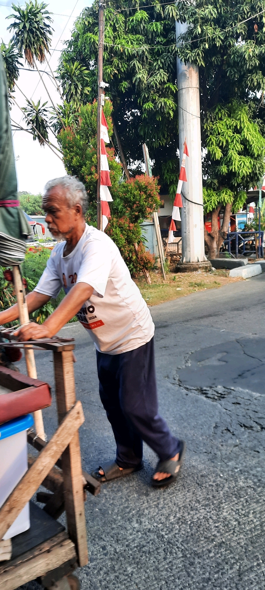 An elderly Indonesian man with gray hair and a beard is pushing a wooden cart on an asphalt road in East Jakarta, Indonesia. He is wearing a light-colored t-shirt with writing on it and dark blue pants. He has a pair of dark sandals on his feet. The cart is made of wood and has a blue and white container on it. Behind the man, there are trees, a utility pole decorated with red and white flags, and some greenery. The road appears to be in a somewhat neglected condition with visible cracks and patches. The lighting suggests it is daytime, possibly late afternoon given the shadows.