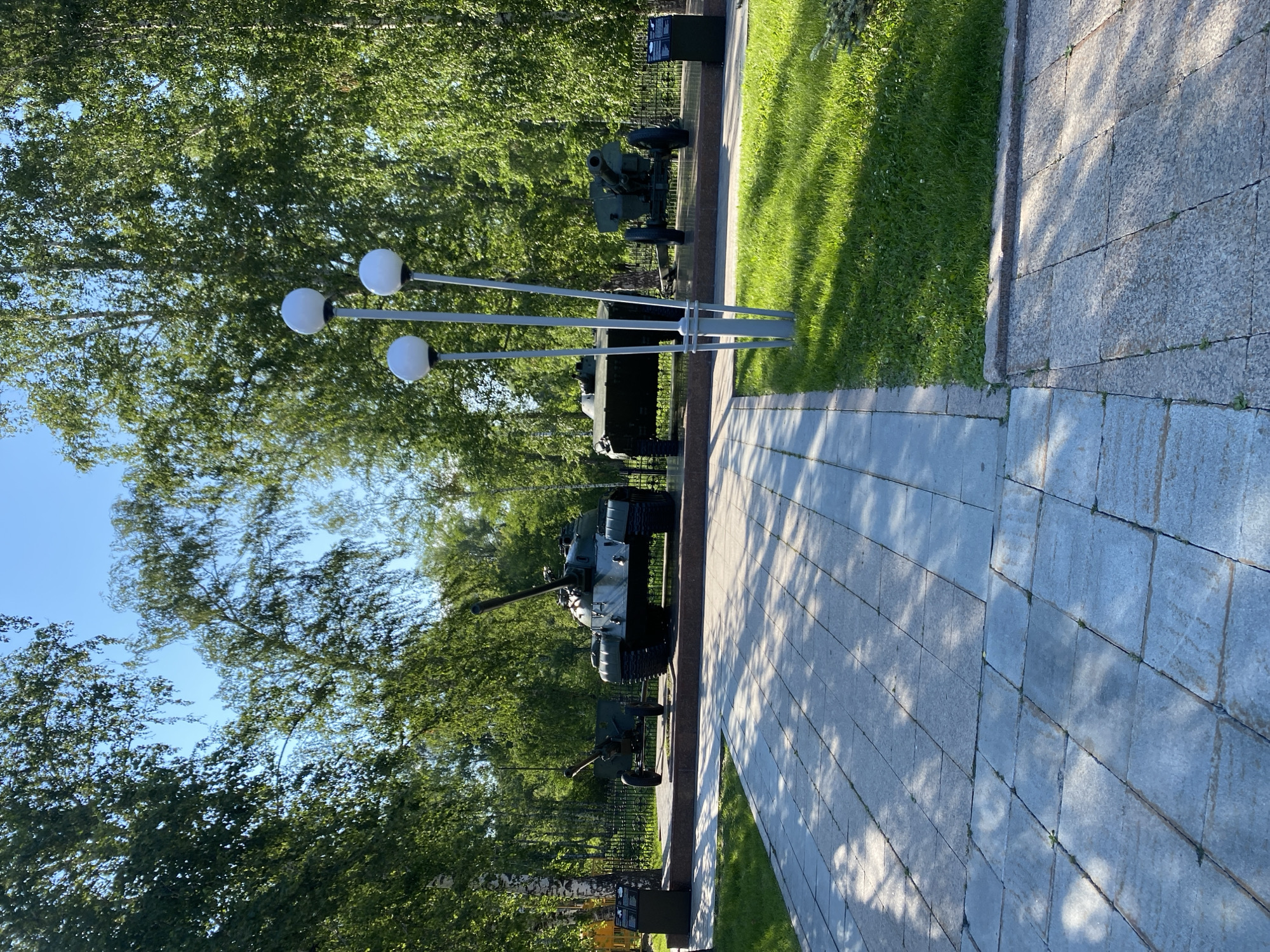 The image shows a military memorial with several static displays of military equipment, including tanks and artillery. The scene is set outdoors with lush green trees and a clear blue sky, suggesting a sunny day. A paved walkway runs alongside a grassy area, with the military displays positioned on raised platforms. A modern-style lamp post with three spherical lights is also visible in the foreground. The equipment appears to be part of a museum or a permanent outdoor exhibition. The overall atmosphere is peaceful and contemplative, likely a place of remembrance. There is no visible text that can be deciphered.