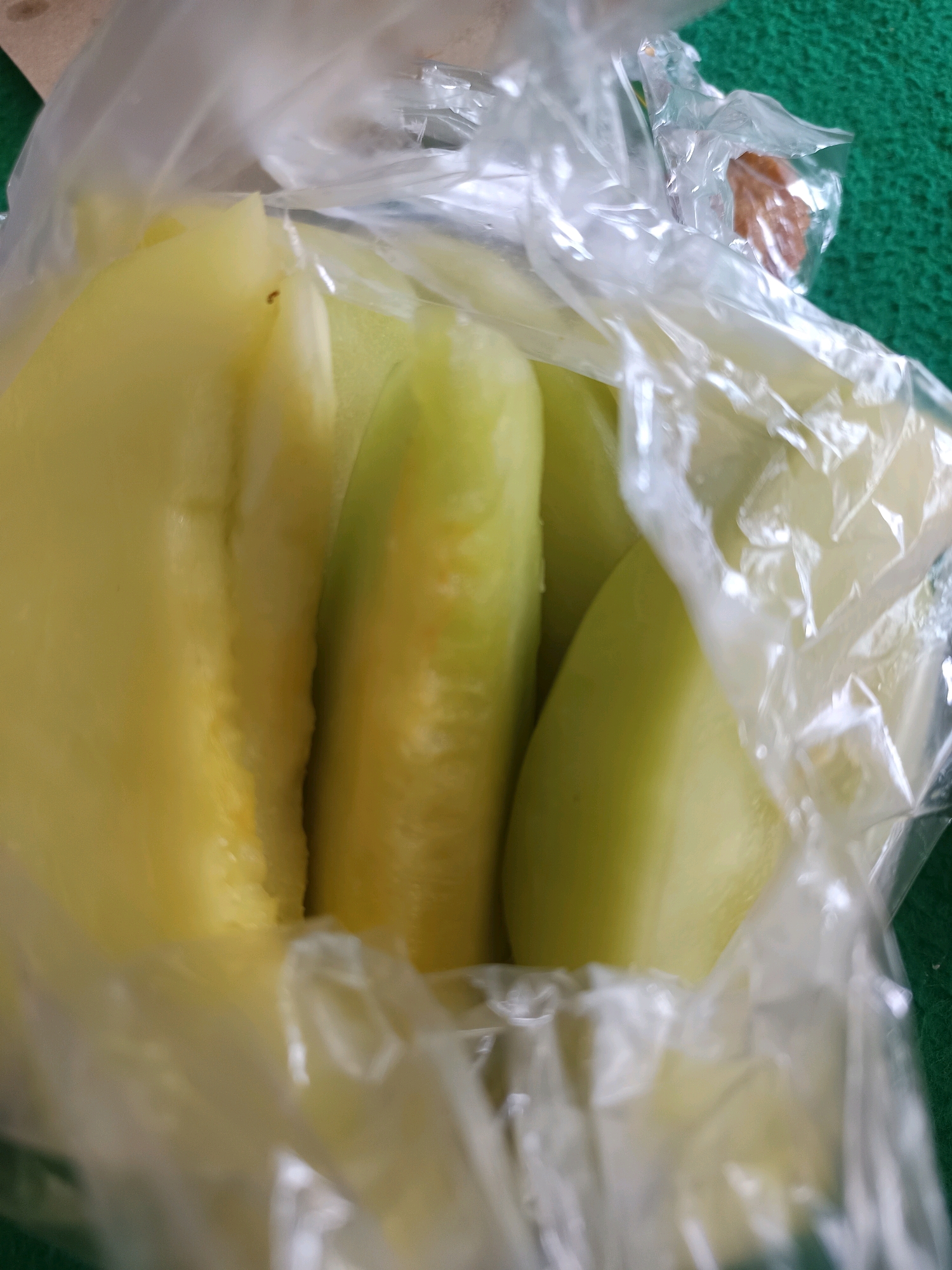 The image shows sliced star fruit in a clear plastic bag, possibly on a green textured surface. The star fruit slices are a pale yellow and appear juicy. A small dark object, possibly an ant, is visible on one of the fruit slices. The background is blurred and out of focus, but a piece of brown material and some green texture can be discerned. The scene appears to be a casual setting, perhaps a market or a home, in Labuhan, Indonesia.
