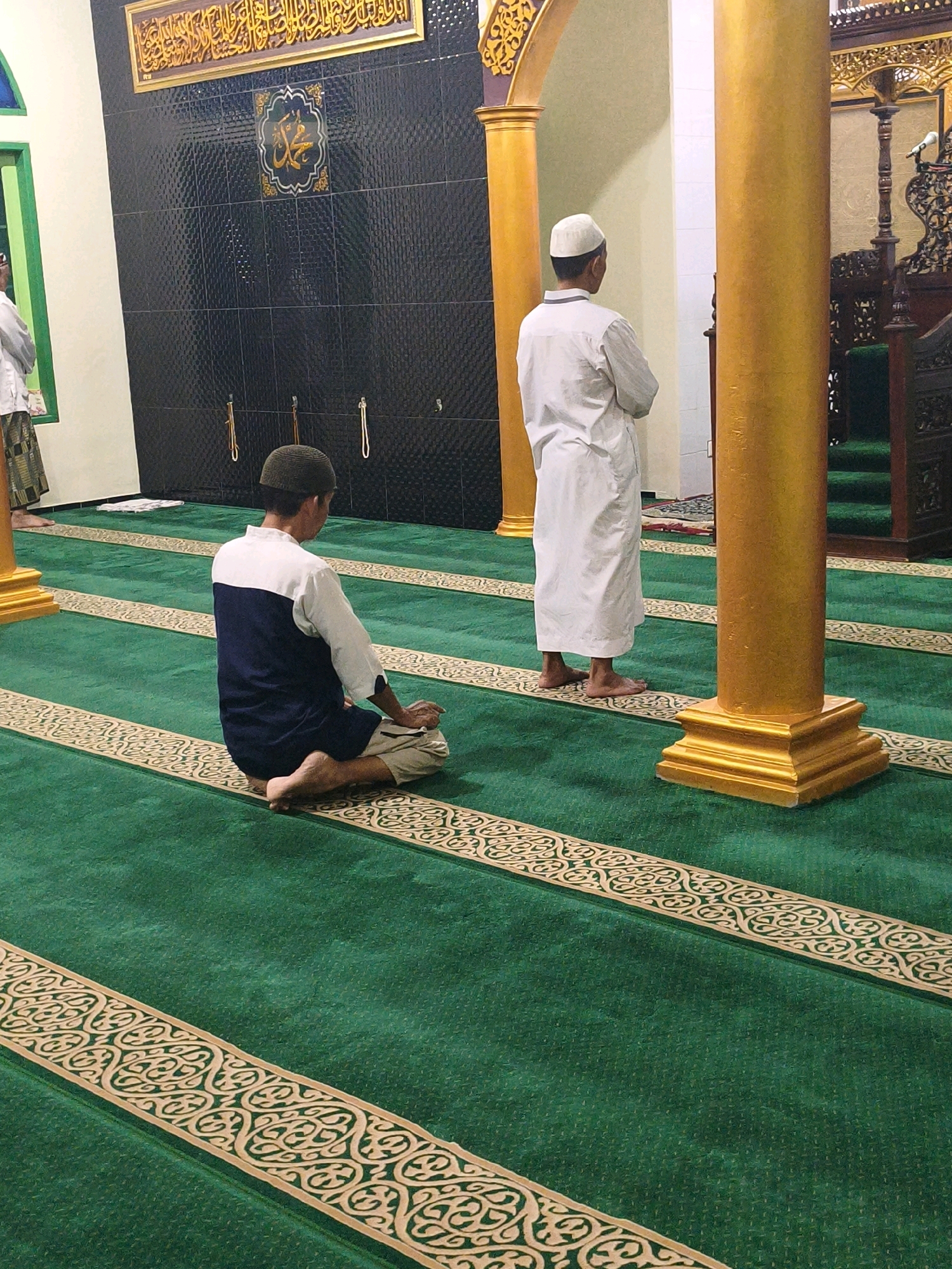 In the city of Samarinda, Indonesia, a mosque interior features green carpeting with intricate gold borders. Two men are present. In the foreground, a man wearing a dark blue and white shirt and tan pants is kneeling on the carpet, facing away from the camera. In the background, another man in a white robe and cap stands facing a wooden pulpit, also with his back to the camera. Golden pillars are visible within the mosque, along with a black tiled wall adorned with Islamic calligraphy.
