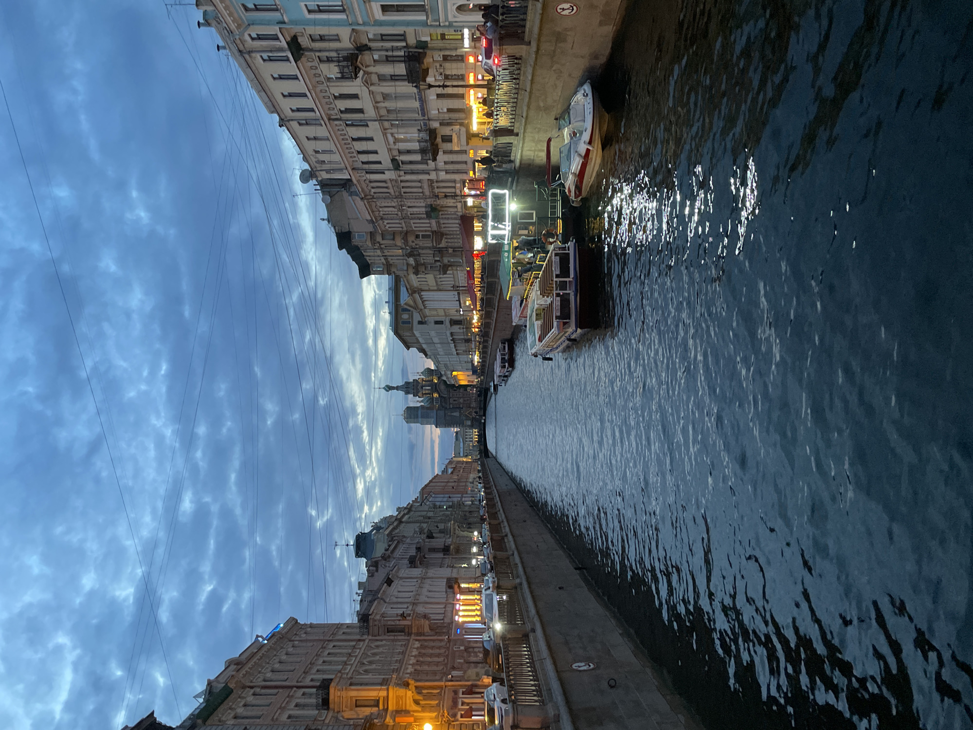 The image captures a twilight scene along a canal in Saint Petersburg, Russia, likely the Griboyedov Canal, given the prominent view of the Church of the Savior on Spilled Blood in the distance. The canal runs centrally through the frame, reflecting the moody, cloudy sky and the lights from the surrounding buildings.
On the left bank, a row of multi-story buildings lines the canal, their facades illuminated by warm, artificial light from shop windows and streetlights. Cars are parked along the sidewalk, and a metal railing separates the walkway from the canal.
On the right bank, similar architecture continues, with a more bustling atmosphere evident. Several boats are docked along the canal, including tour boats with people visible on deck, and smaller pleasure boats. Some of these boats are brightly lit. People are walking along the embankment, and the lights of cafes and shops are visible.
The canal water is dark and choppy, with streaks of light reflecting off its surface from the illuminated boats and buildings. The sky overhead is a deep blue, filled with dramatic, textured clouds, suggesting a transition from day to night. Overhead, electrical wires crisscross the sky, adding to the urban feel of the scene. In the far distance, the iconic onion domes of the Church of the Savior on Spilled Blood are visible, silhouetted against the fading light.