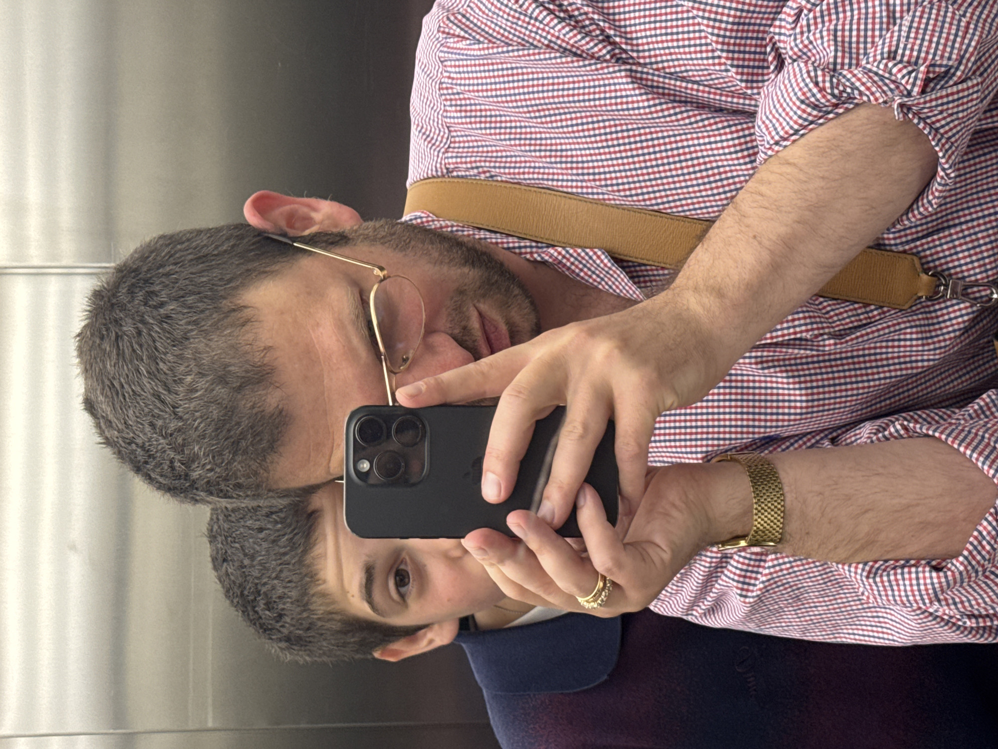 A person takes a selfie in a mirror in an elevator. The person wears a checkered shirt, glasses, a gold watch, and a gold ring. They are holding a black smartphone with three camera lenses. The reflection shows another person in a dark blue collared shirt. The elevator has metal walls. The location is not discernible to be Rome, Italy from this image alone.