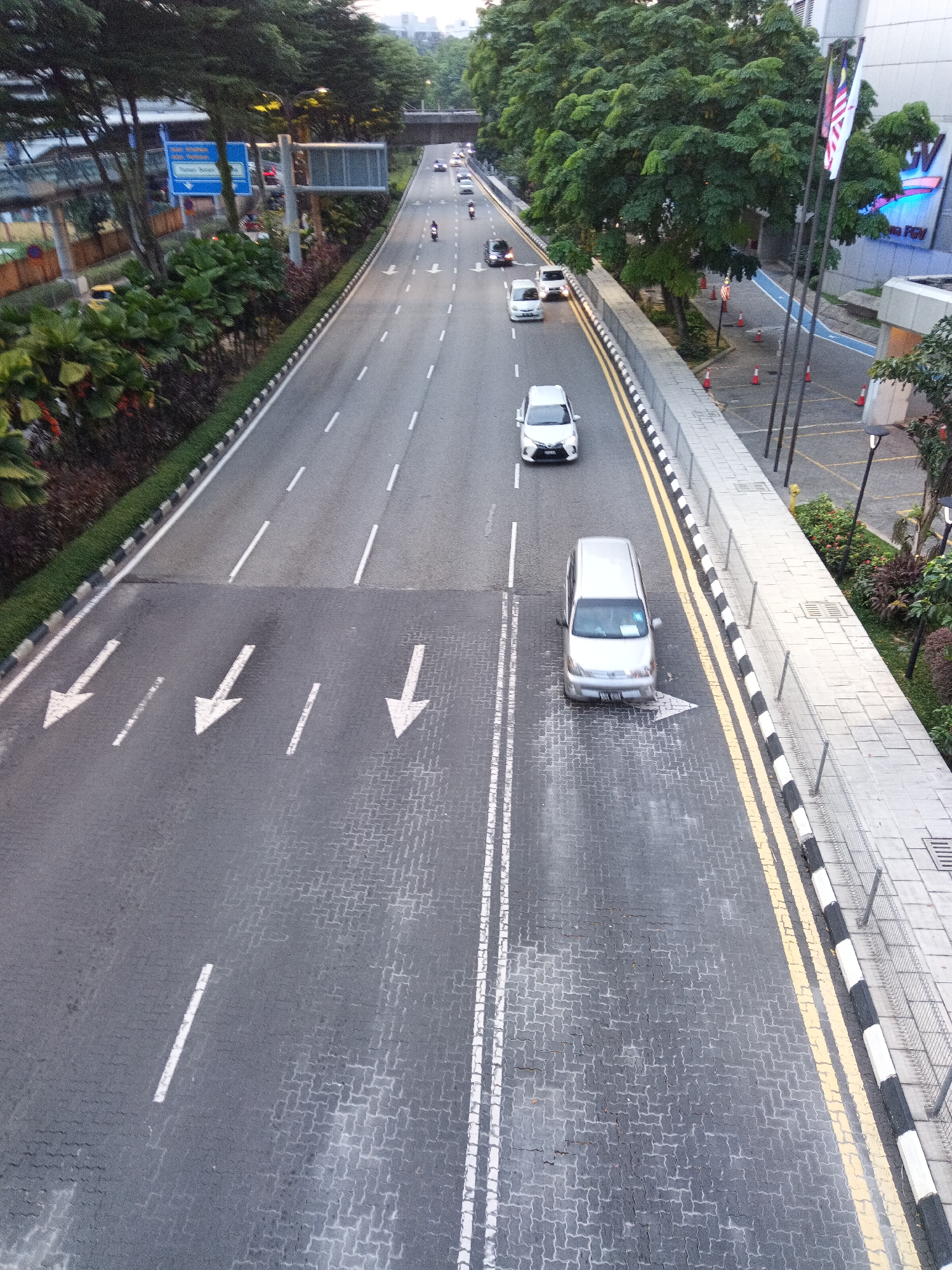 This is a high-angle, street-level, full shot of a multi-lane road in Kuala Lumpur, Malaysia. The road is paved with interlocking bricks and asphalt. White arrows painted on the road indicate traffic direction. Several vehicles are visible on the road, including white cars and a silver van. Green trees line the sides of the road, and there are buildings and streetlights visible in the background. The scene appears to be during daylight hours, likely early morning or late afternoon, given the soft lighting. The overall impression is one of urban infrastructure and daily life in a Southeast Asian city.