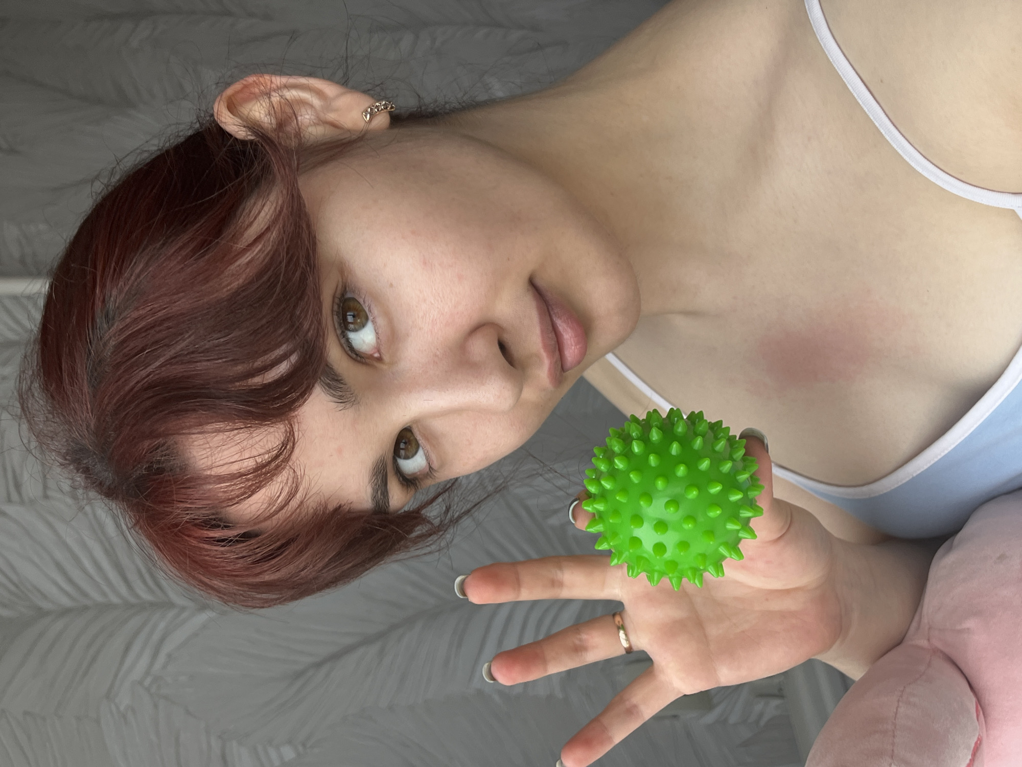 A young woman with dark red hair and fair skin is holding a bright green spiky ball in her right hand. She is wearing a light blue tank top with white straps. A ring is visible on her right ring finger. There is a reddish mark on her chest. A portion of a pink plush toy is visible in the lower right corner of the image. The background is a light gray color with a subtle patterned texture. The image is rotated slightly clockwise. It is not possible to determine the location of the image to be in Neftekamsk, Russia, without additional information.
