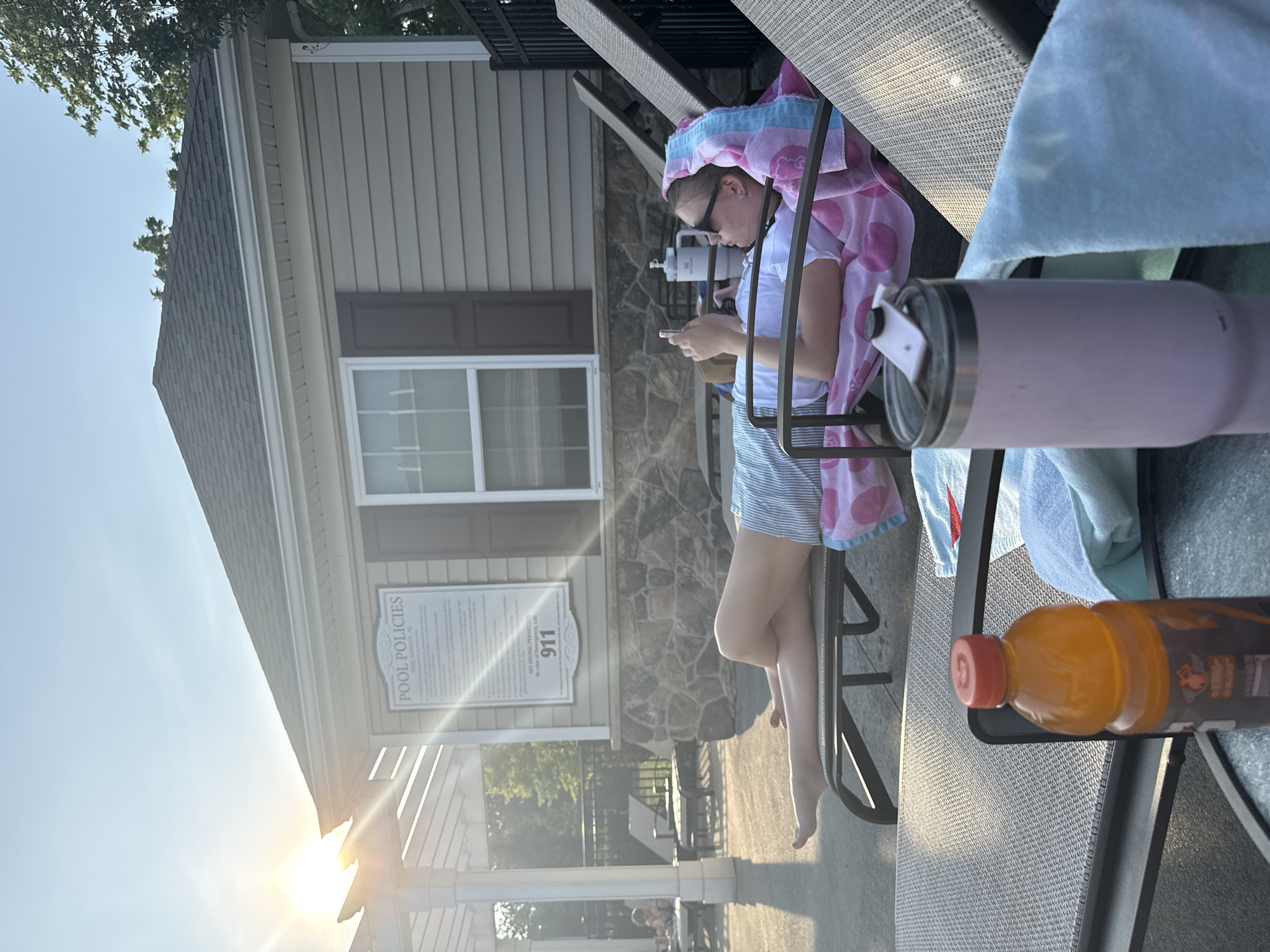 A young person with blonde hair, wearing sunglasses and a blue and white striped skirt, lies on a lounge chair by a pool. They are looking at their phone, with a pink and blue towel draped over their head and shoulders. The sun is setting, casting a warm glow on the scene. A building with "POOL POLICIES" signage is visible in the background. In the foreground, a bottle of orange liquid and a pink tumbler sit on a table. The location is the pool area of a community in Allen Township, United States of America.