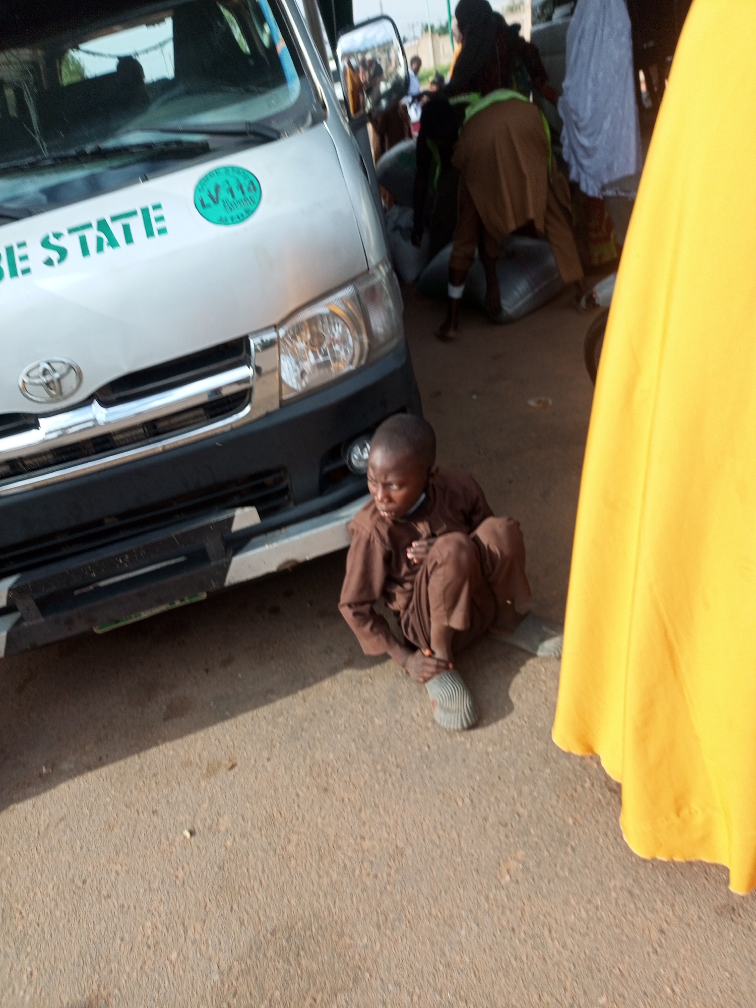 In Gombe, Nigeria, a young boy, likely a child laborer, sits on the ground next to a silver van. He wears a brown tunic and trousers and appears to be barefoot, or wearing very thin socks. His posture suggests weariness, with his knees drawn up to his chest and his hands resting on his legs. The van, emblazoned with "BE STATE" in green lettering and a green circular emblem with "LV114" inside, is positioned in the foreground. In the background, other individuals are visible, some wearing traditional clothing and headscarves. They are engaged in the activity of loading or unloading large sacks, suggesting a busy market or transportation hub. The presence of these sacks and the boy's attire imply a context of labor or commerce. The lighting suggests it is daytime, possibly late morning or early afternoon, with the sun casting shadows. The ground is paved, indicating an urban or semi-urban setting. The overall scene conveys a sense of daily life and economic activity in Nigeria, with the child's solitary presence by the van hinting at his role in this environment.