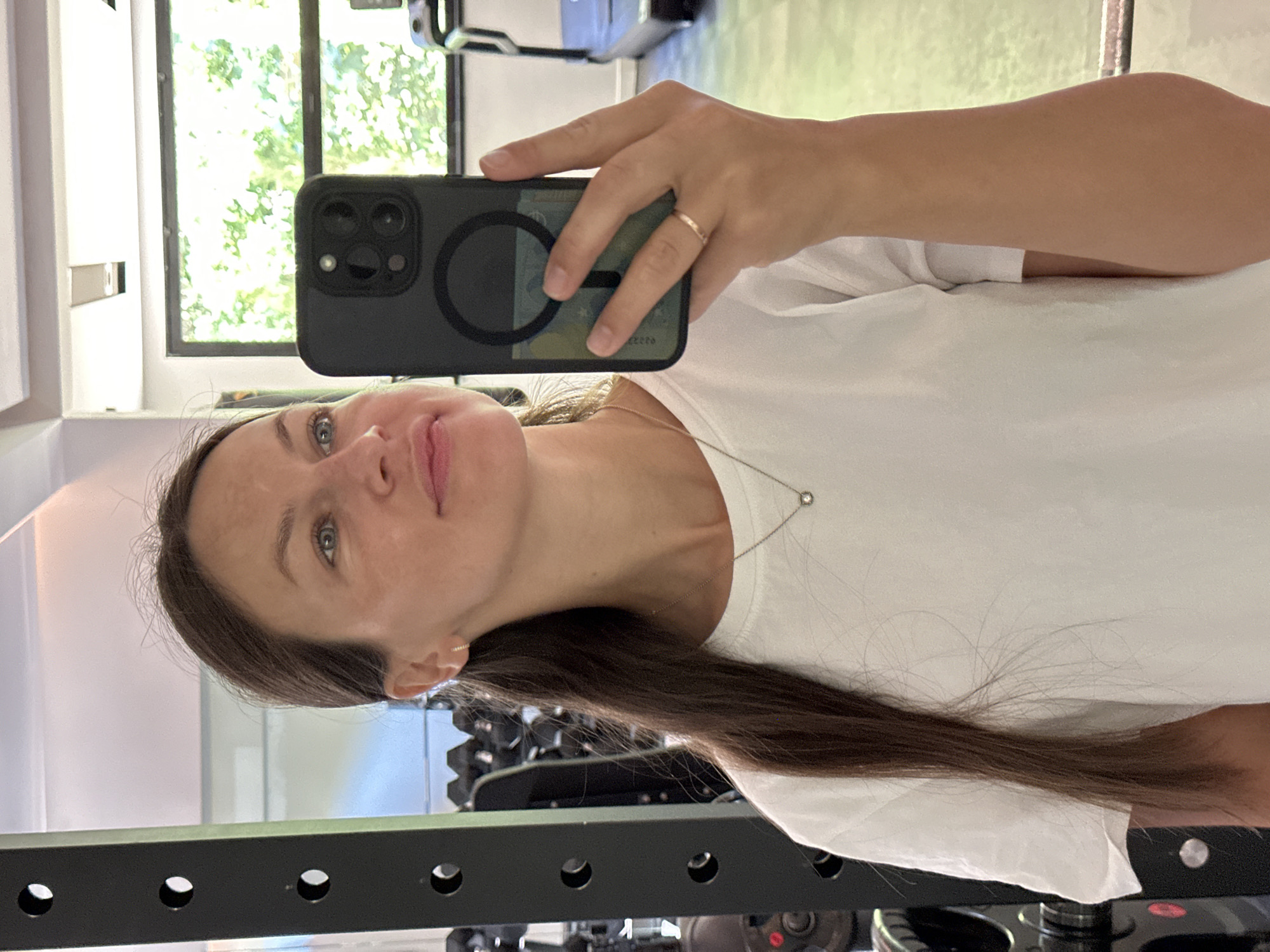 A woman is taking a mirror selfie in a gym. She is wearing a white t-shirt and a thin necklace with a small pendant. She has long brown hair pulled back. Her right hand is holding a black phone case with a green card visible inside. A ring is on her left ring finger. In the background, a weight rack is on the left, and a treadmill is visible on the right through a large window. The gym appears to be located in Bečići, Montenegro.